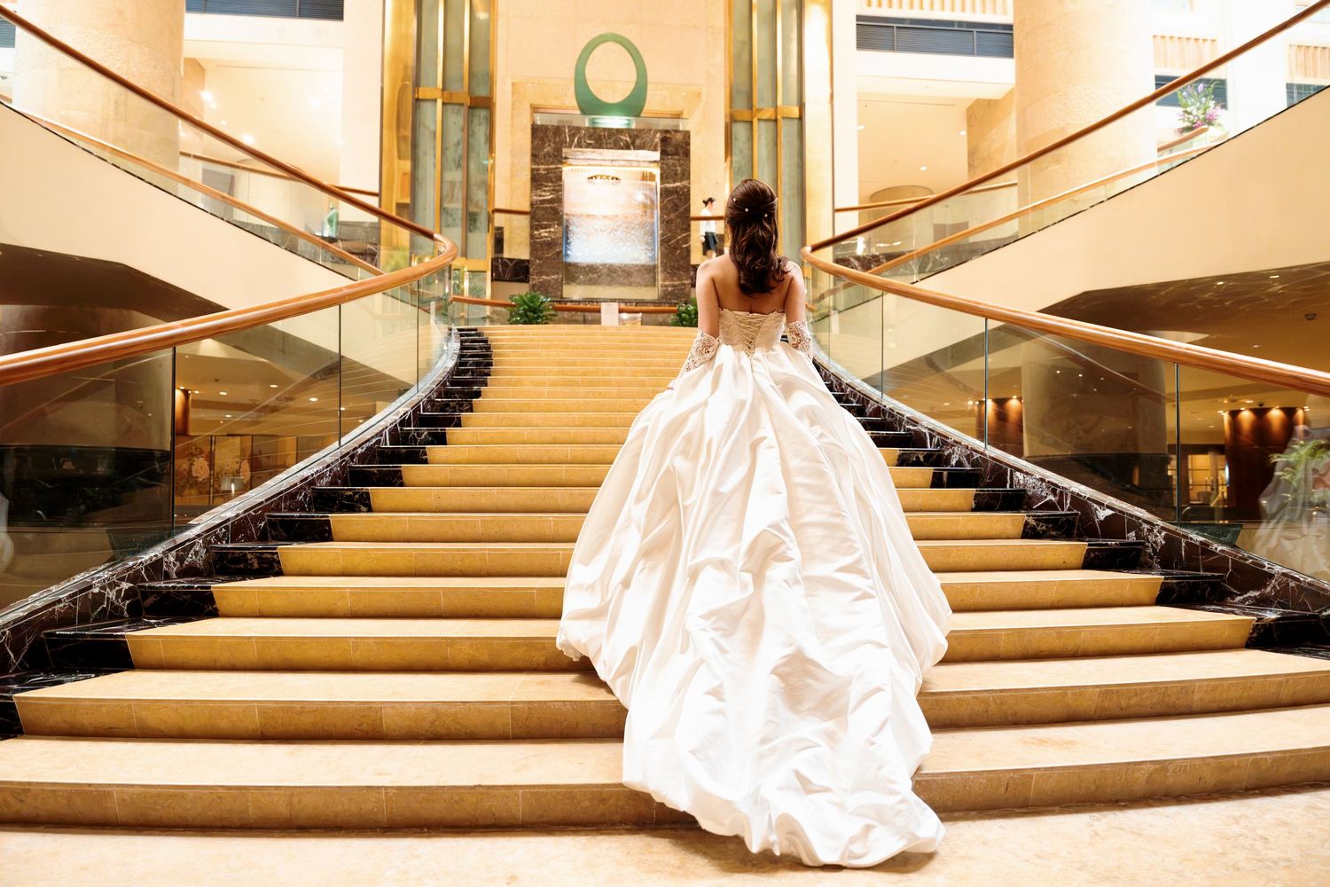Bride in a flowing white ball gown with an open back climbs a grand curved staircase in an elegant hotel lobby.