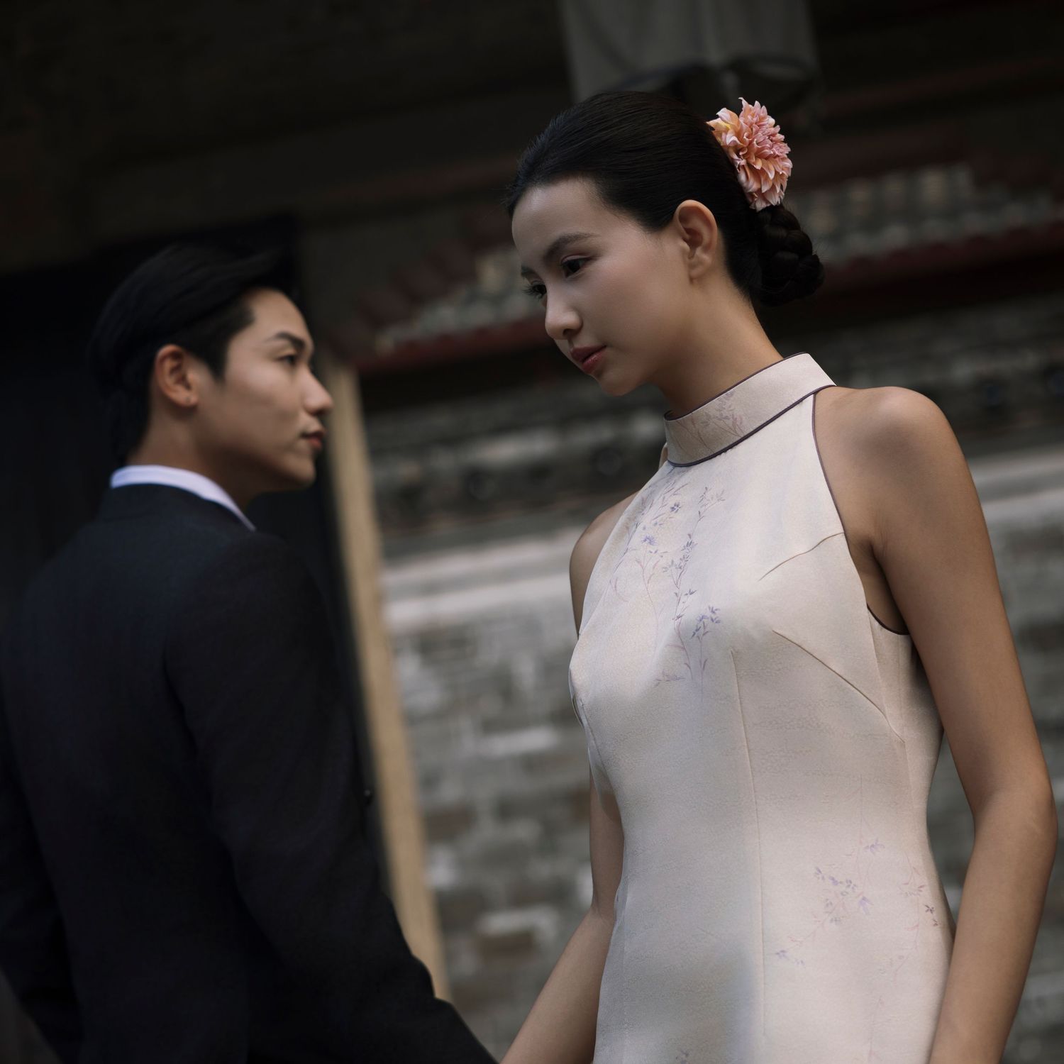 A couple in elegant attire, woman in white qipao with floral hair accessory, man in black suit, in a traditional setting.