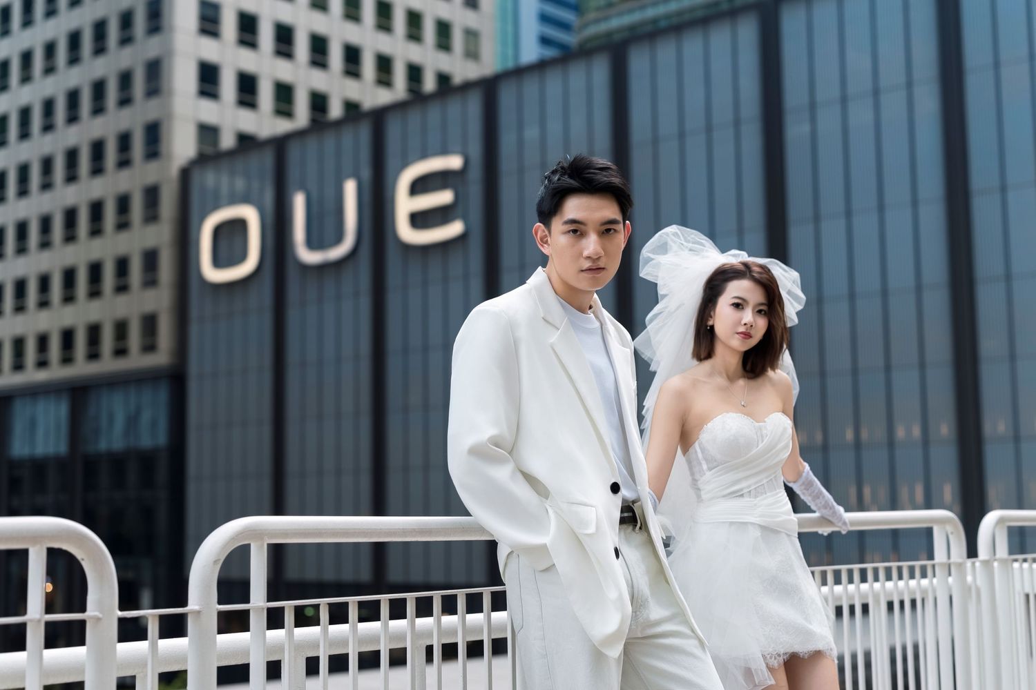 Stylish couple in white wedding attire pose confidently in front of the OUE building in the city.