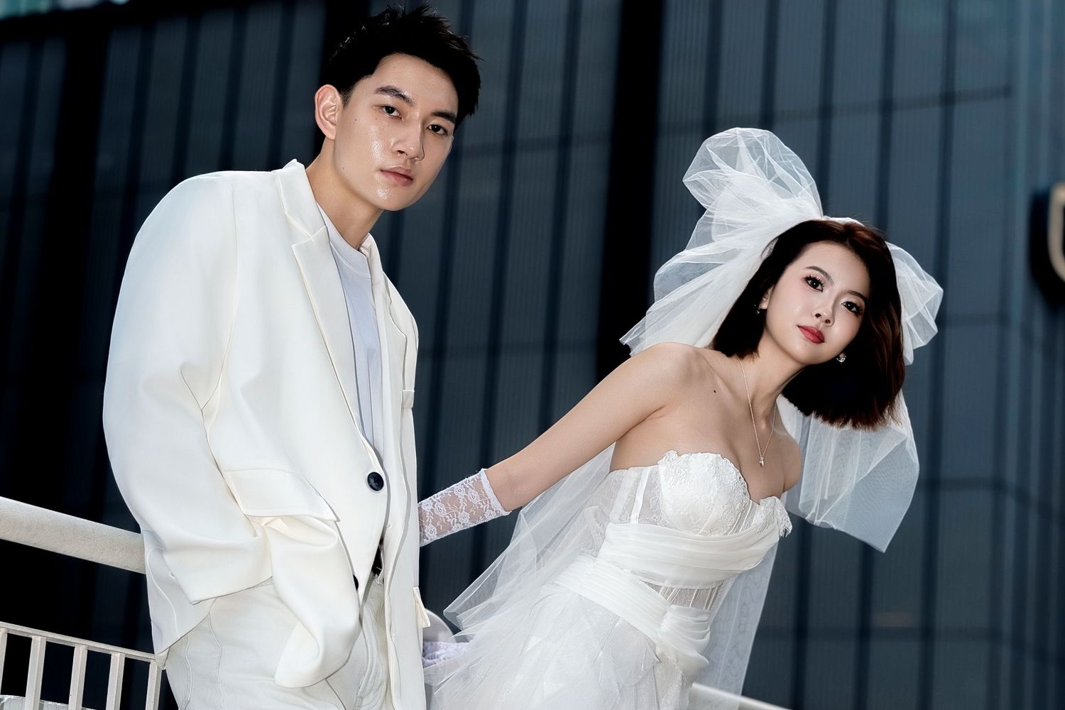 Elegant Asian couple in matching white wedding outfits posing against a modern urban background.