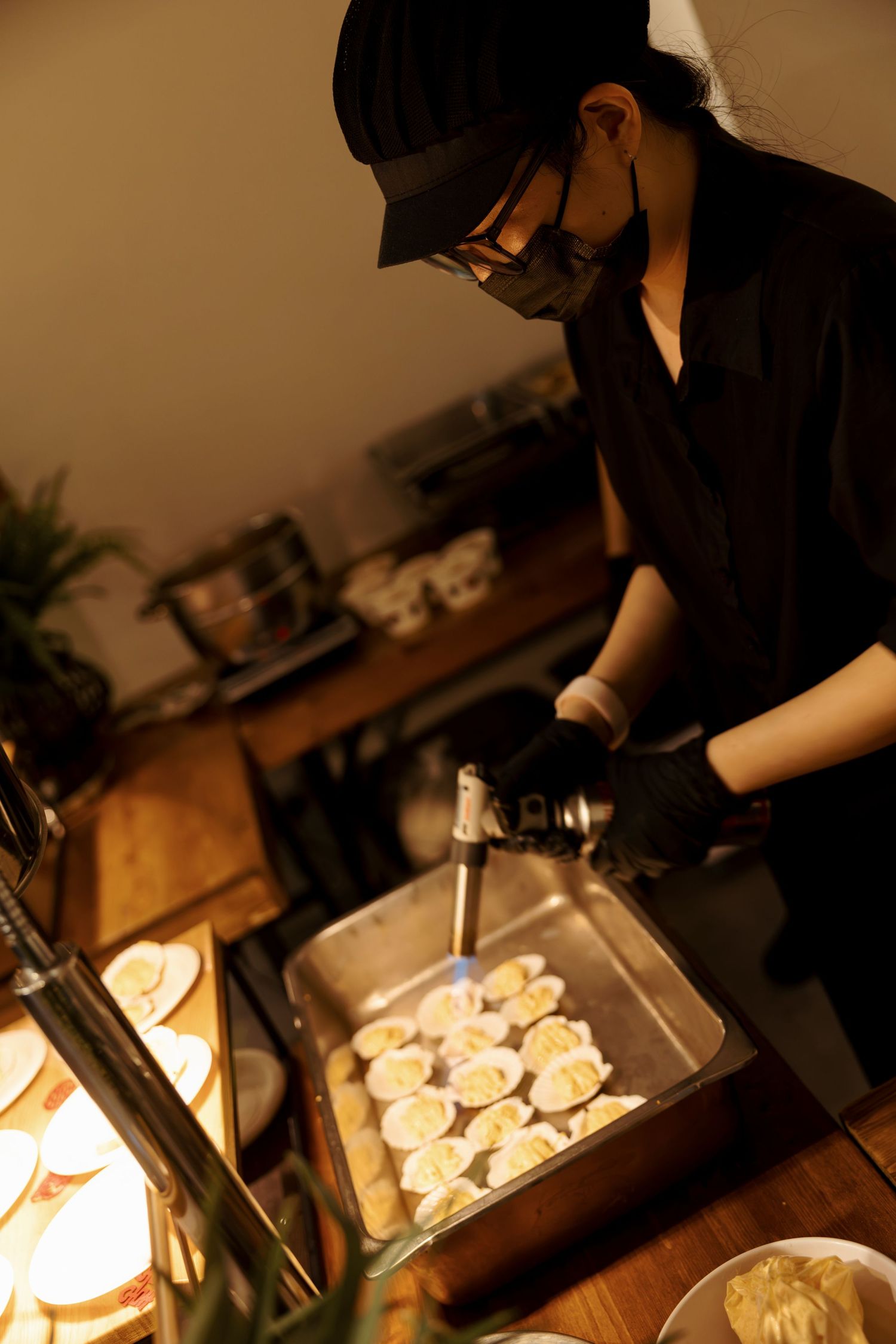 Chef in black uniform using a torch to prepare dumplings in a metal tray at a dimly lit restaurant kitchen.