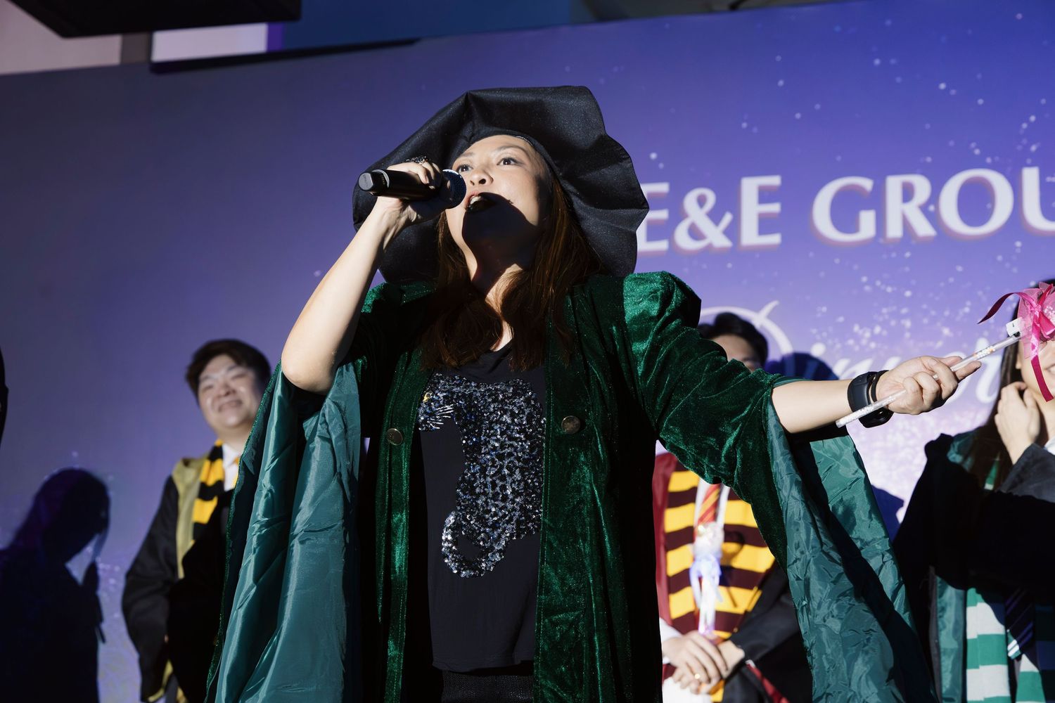 Performer in green graduation robe singing on stage at C&E Group event with confetti.