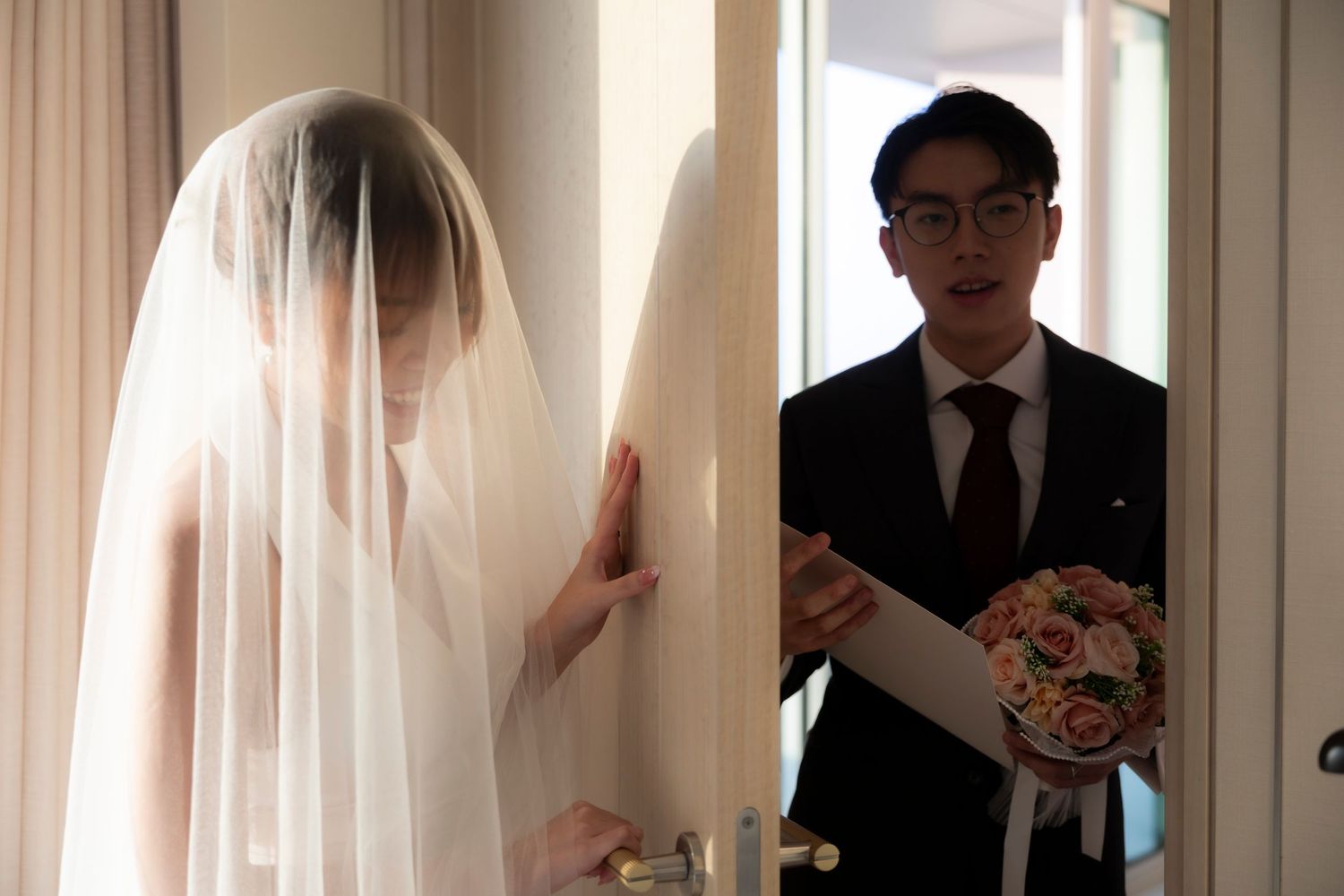 Bride in a sheer white veil opens a door as a groom in a dark suit and glasses waits outside holding a pink floral bouquet.