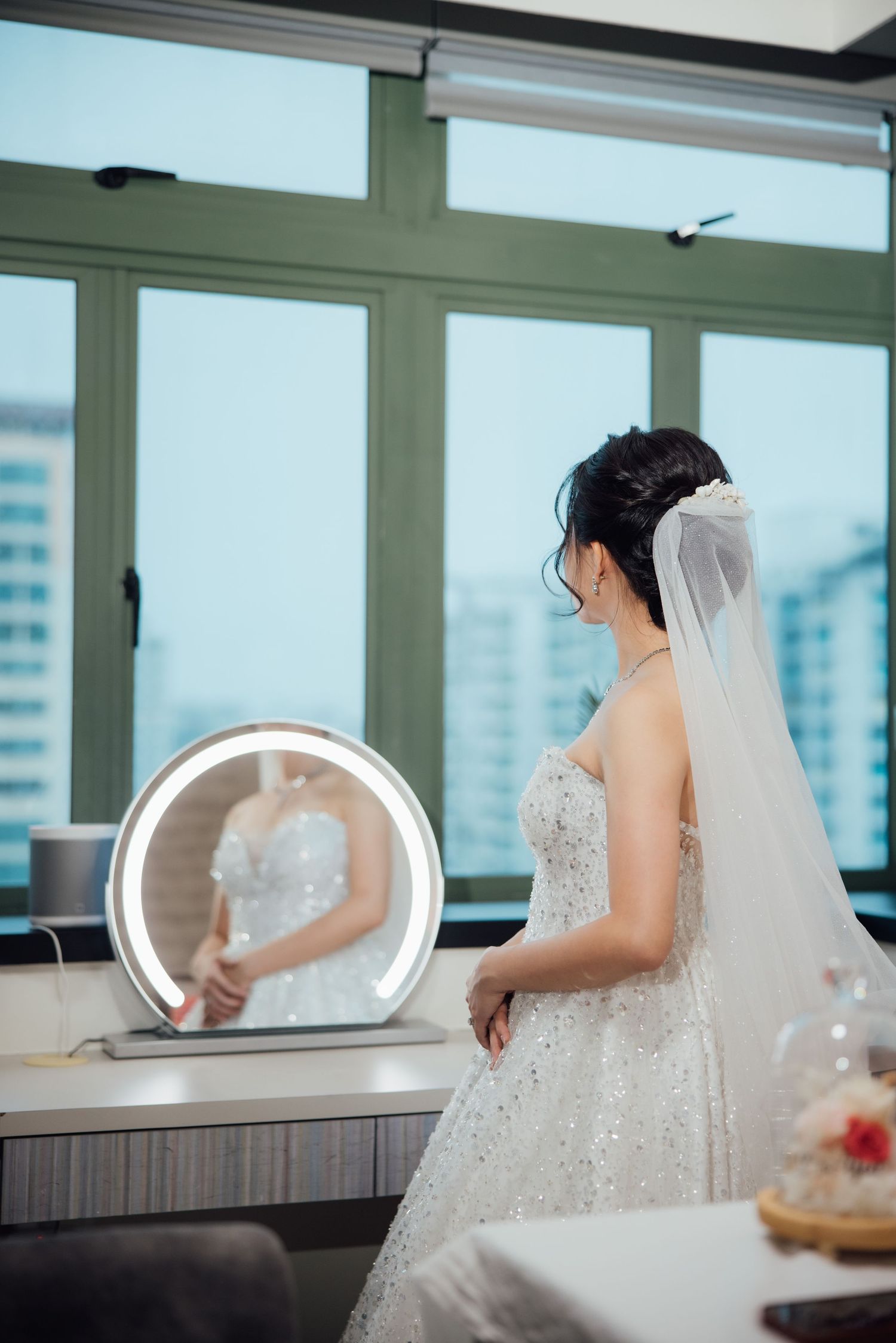 Bride in a sparkling beaded wedding gown and long veil admires her reflection in a lit vanity mirror beside tall windows.