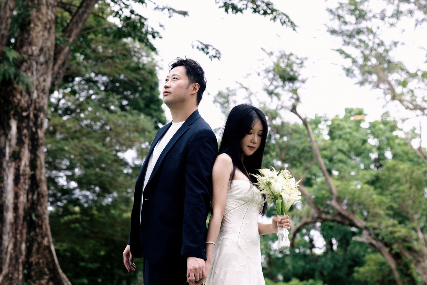 Asian couple in wedding attire standing back-to-back outdoors, bride holding white flowers, groom in black suit, lush green trees.