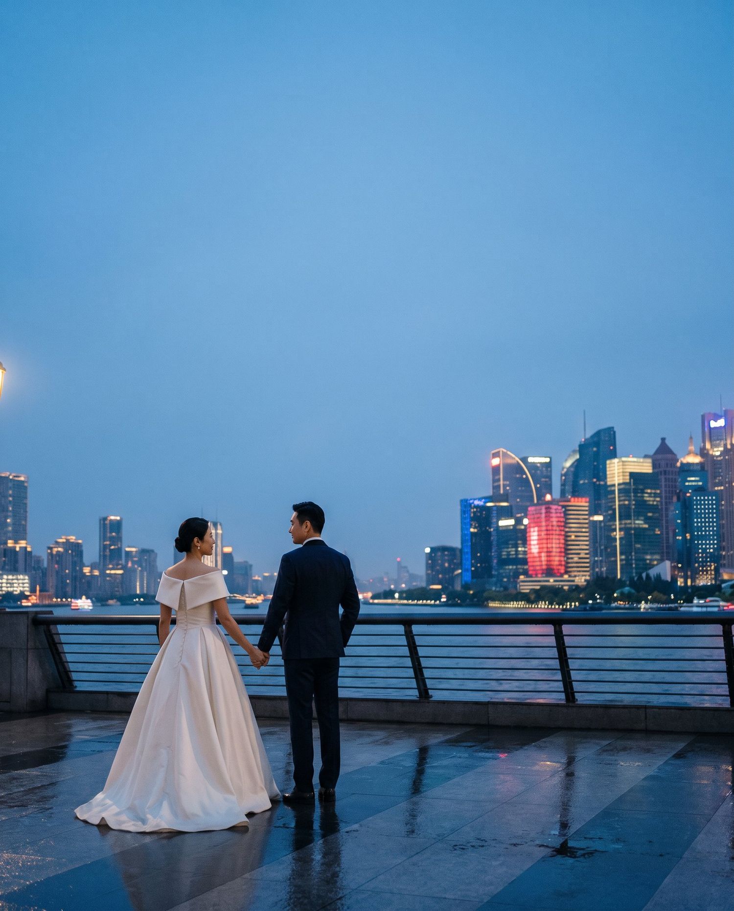 Bride and groom holding hands on a waterfront promenade with a glowing city skyline at dusk.