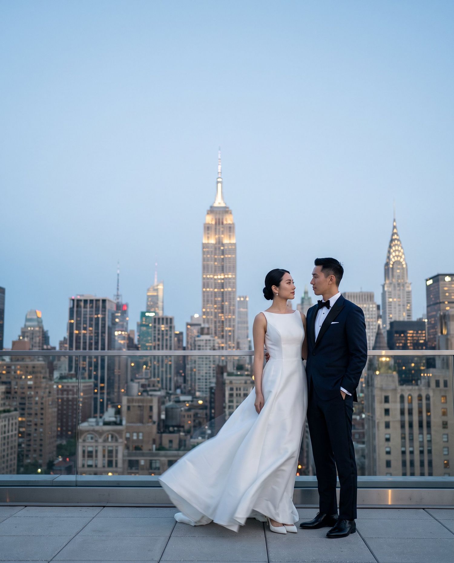 Bride and groom pose on NYC rooftop with Empire State Building at dusk in elegant wedding attire.