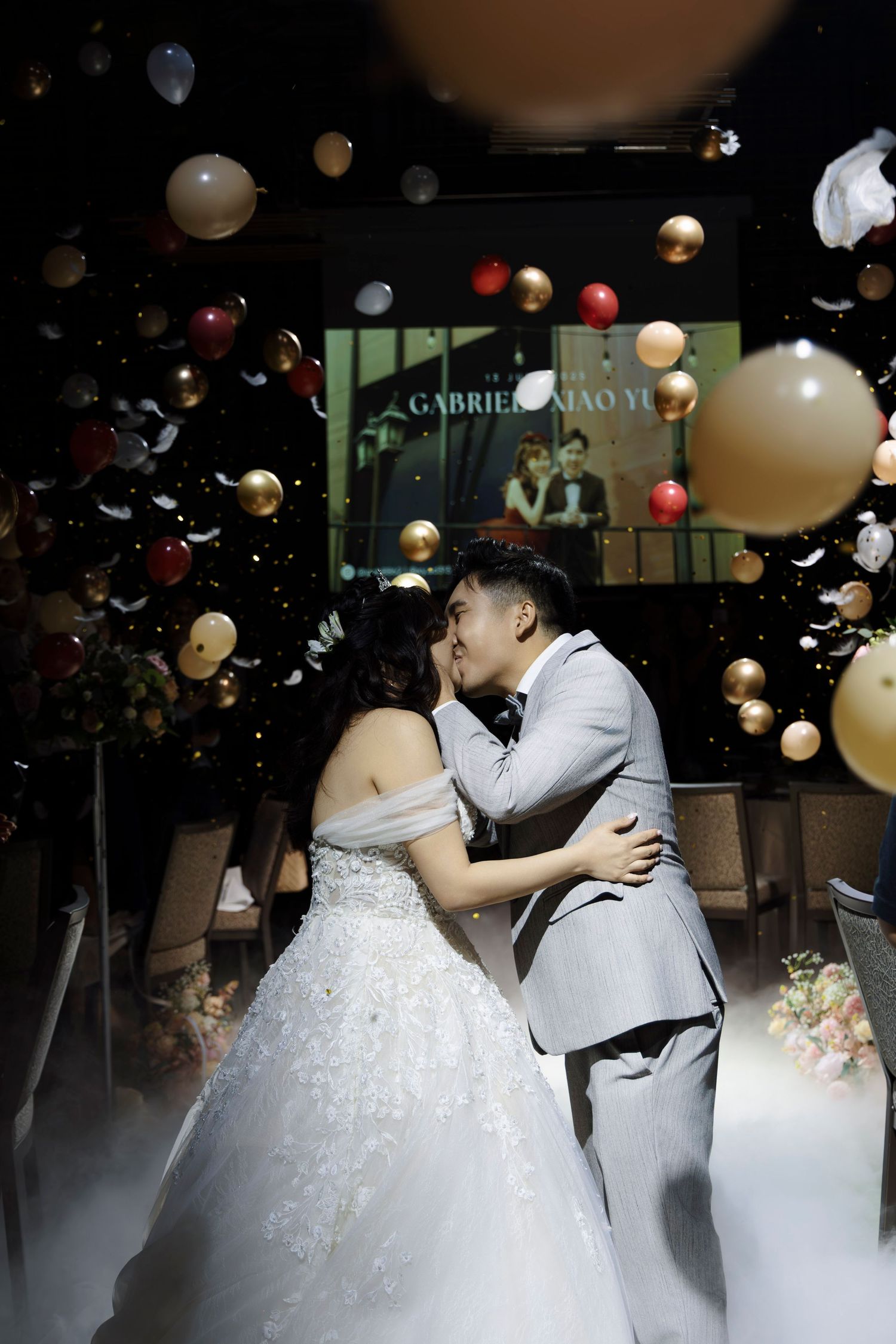 Bride and groom share first kiss surrounded by floating balloons and confetti at wedding reception.