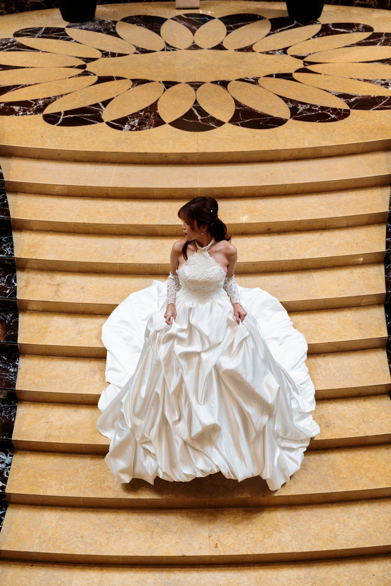 Bride in a white satin ball gown with lace sleeves stands on wide marble steps beneath a floral medallion floor design.
