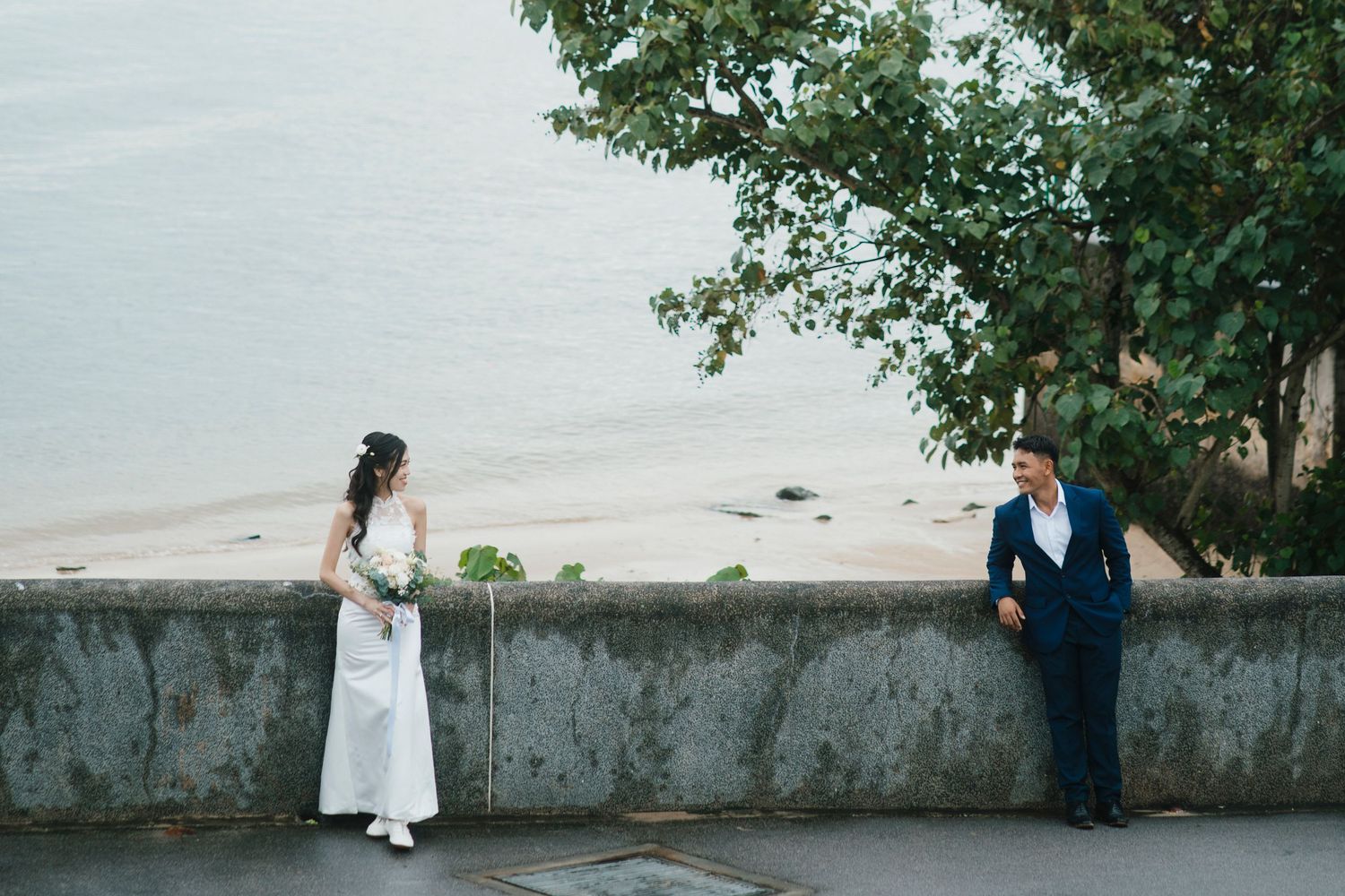 Bride in white dress and groom in blue suit pose by a seaside wall with a sandy beach backdrop.