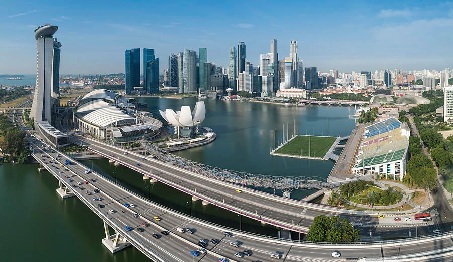 Aerial panoramic view of Singapore skyline with Marina Bay, highways, and modern skyscrapers under a clear blue sky.