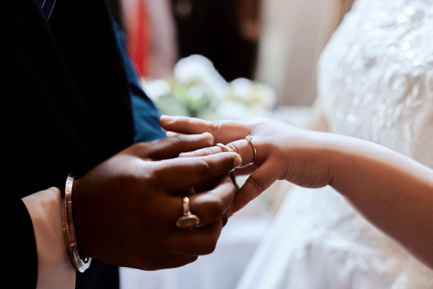 Close-up of interracial couple exchanging wedding rings during marriage ceremony, bride in white gown.