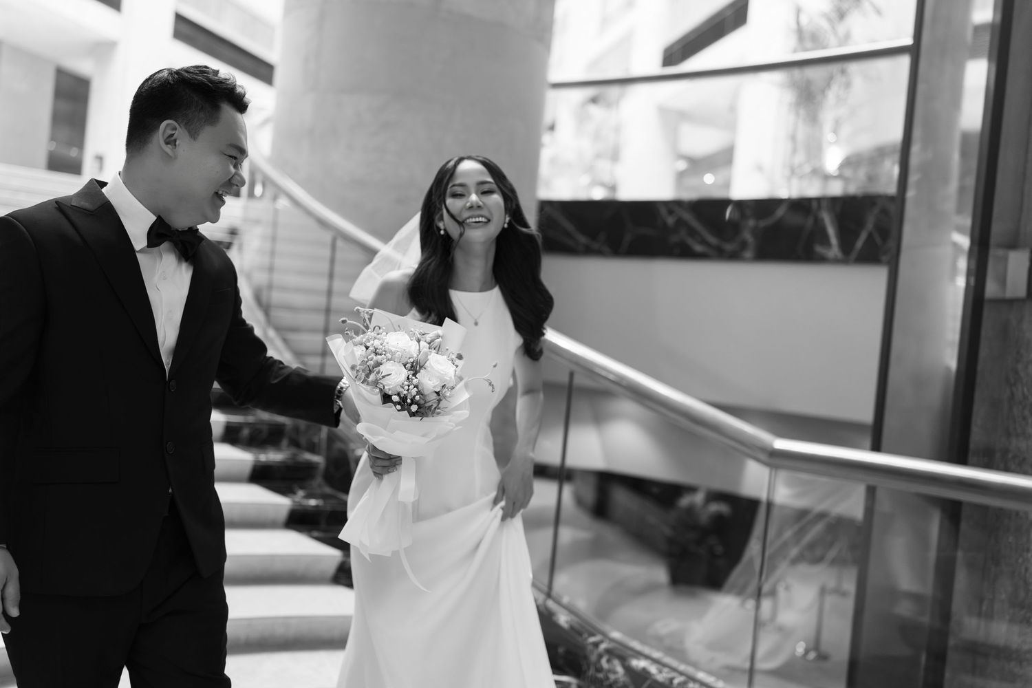 Black and white photo of smiling bride walking with groom holding floral bouquet indoors.