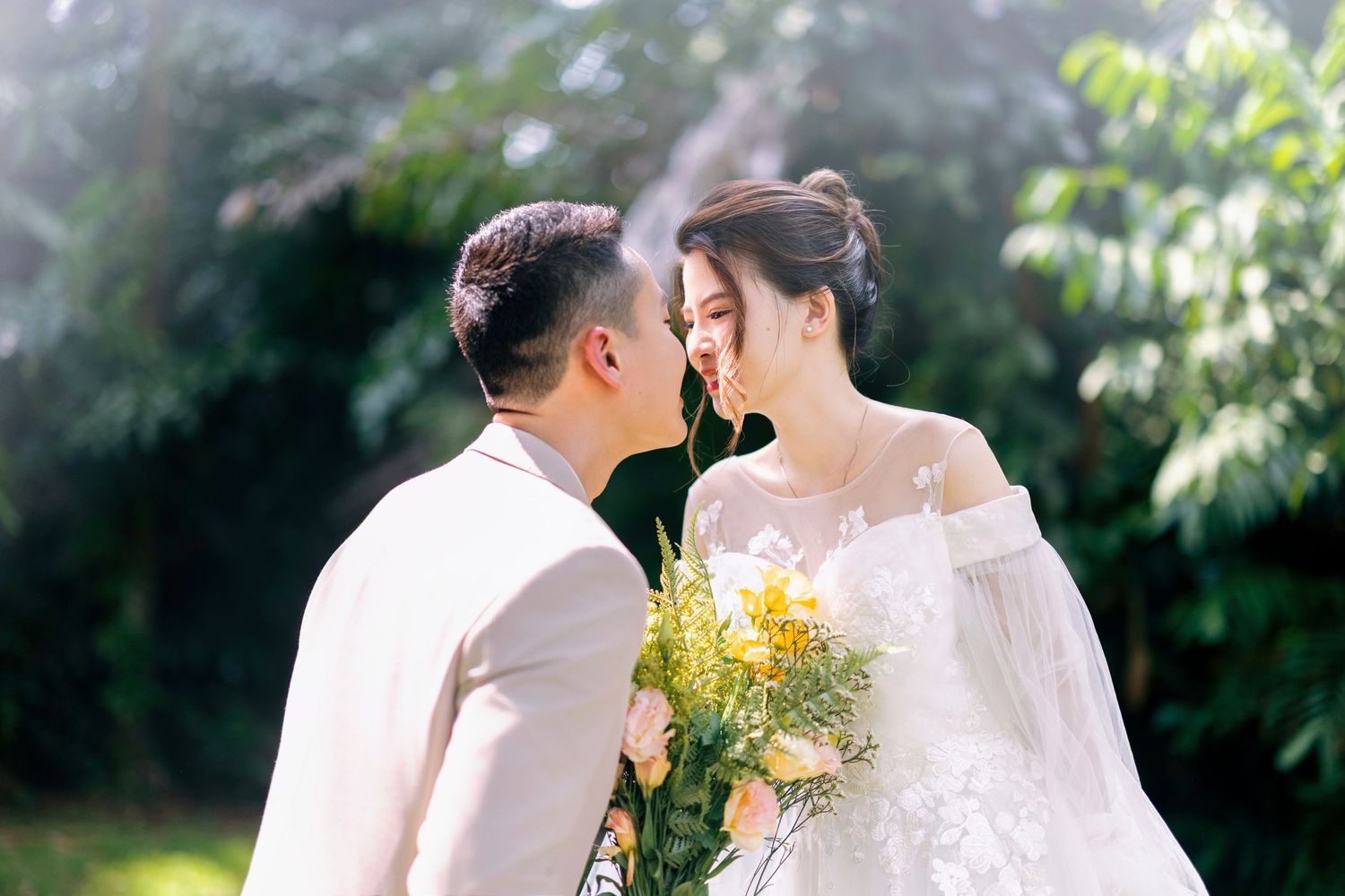 A couple in wedding attire shares a romantic moment outdoors, bride holding yellow flowers, surrounded by lush greenery.