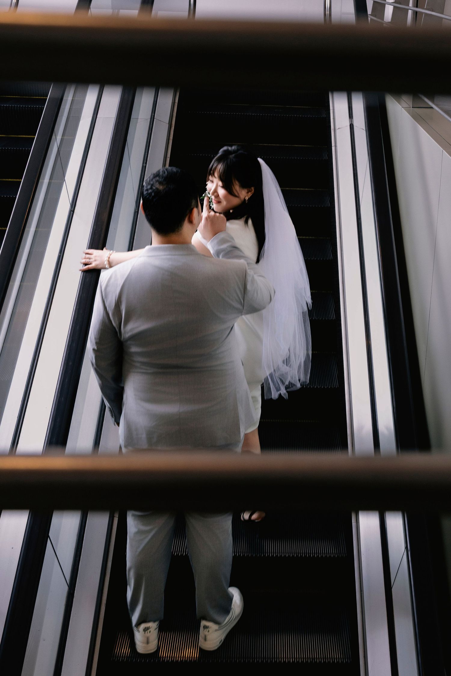 Bride and groom sharing a tender moment on an escalator, bride in white gown and veil, groom in gray suit.