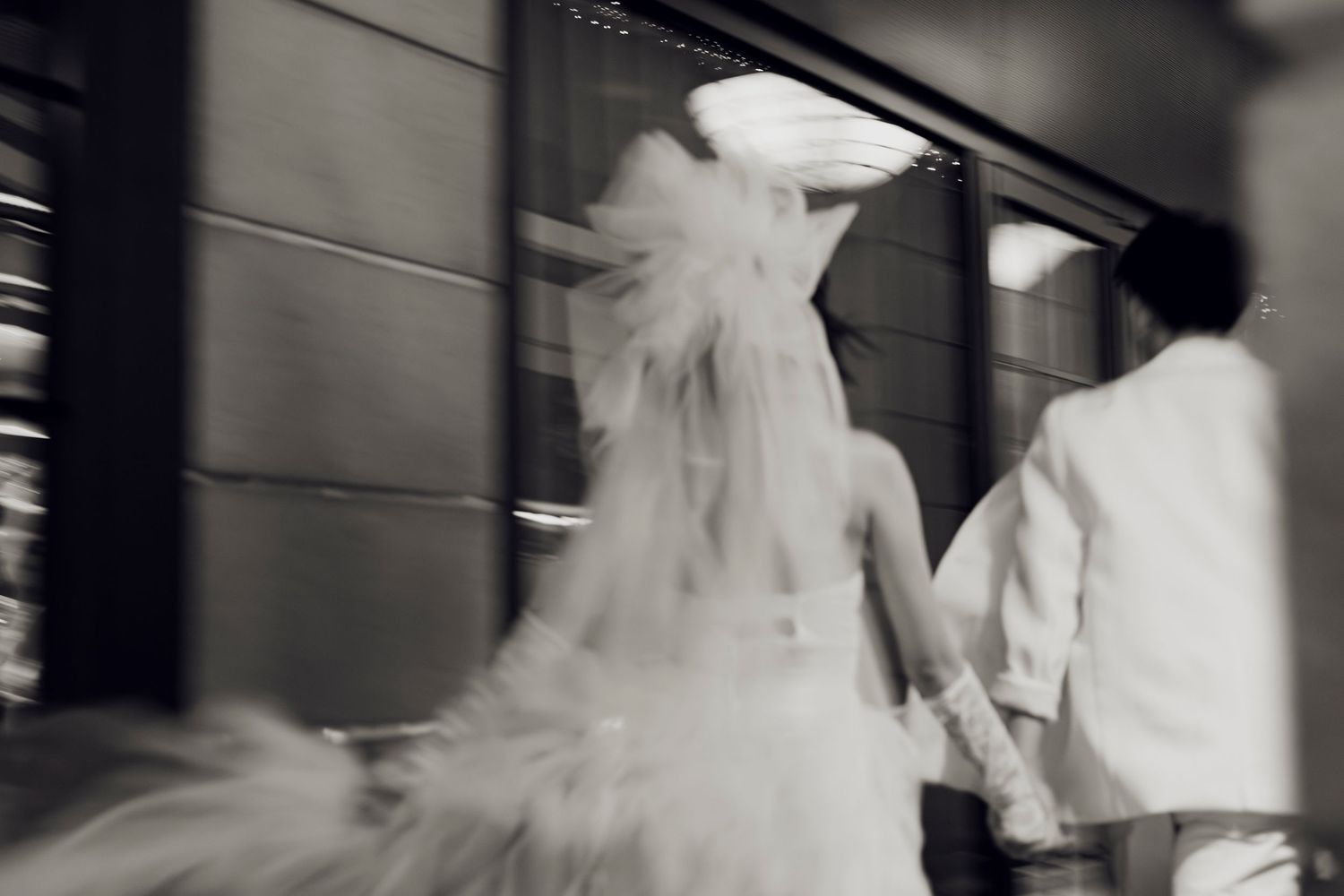 Blurred black and white photo of a bride in a flowing wedding gown and groom holding hands walking together.