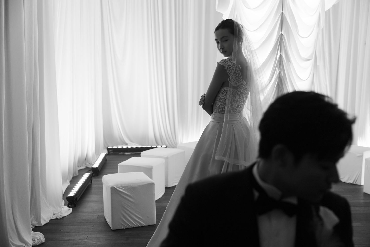 Black and white wedding portrait of a bride in a veil glancing over her shoulder beside a groom in a bow tie, with sheer curtains behind.
