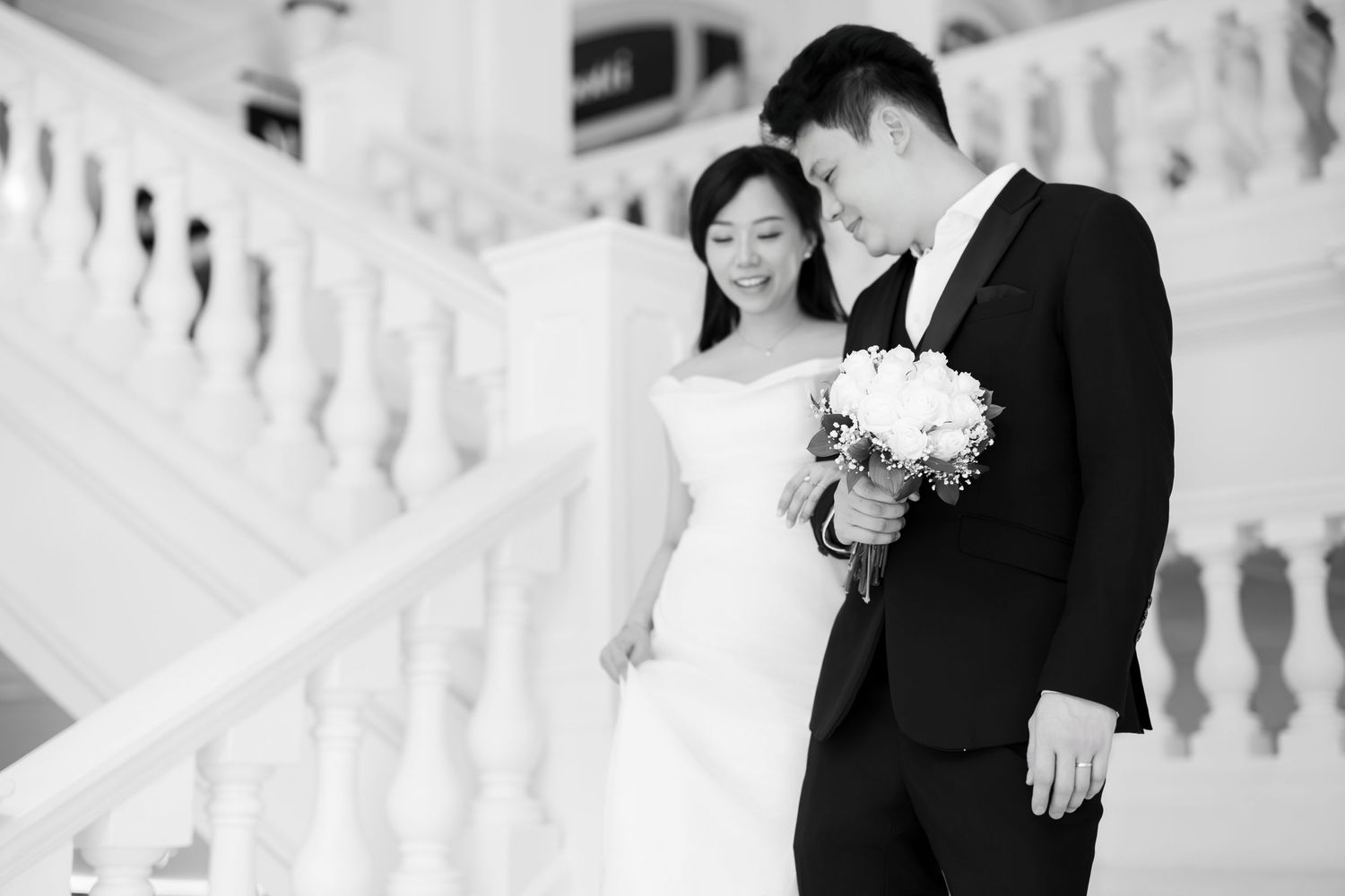 Black and white photo of an Asian bride and groom posing on elegant white staircase with bouquet.