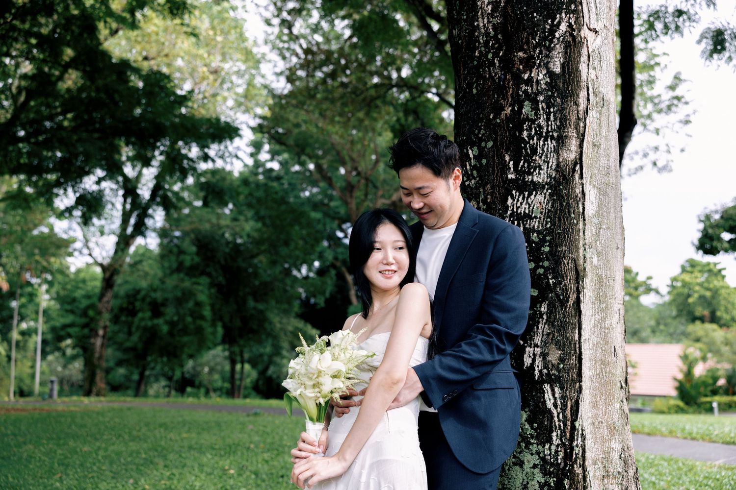 Couple in wedding attire embracing by a tree in a lush green park, bride holding a bouquet.