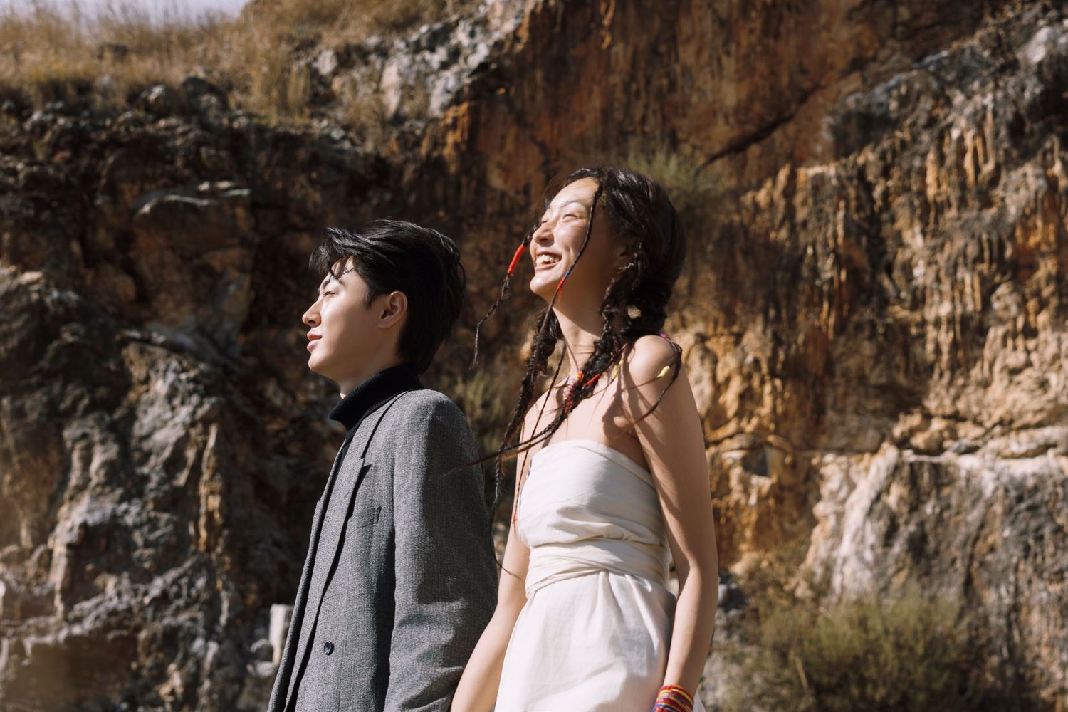 Couple in wedding attire standing before rocky cliffs, woman in white dress laughing joyfully outdoors.