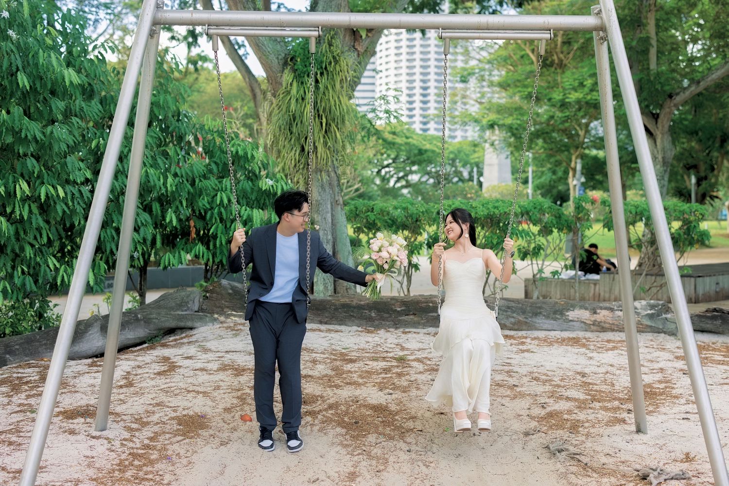 Couple in wedding attire holding hands and smiling at each other near a swing set in a lush green park.