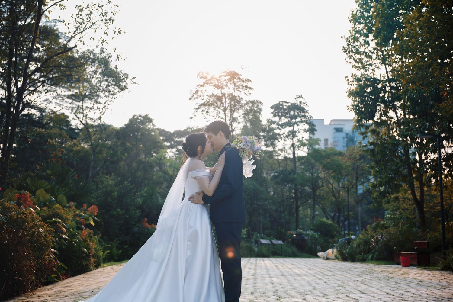 A bride and groom share a romantic moment outdoors, surrounded by lush green trees with soft sunlight in the background.