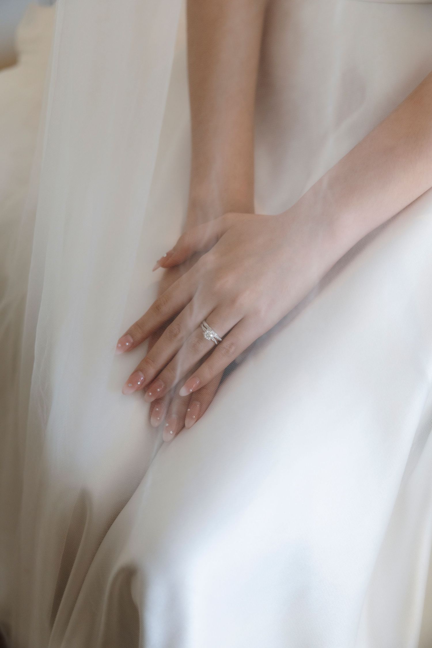 Bride's hands with pink manicure and sparkling diamond engagement ring resting gently on her flowing white wedding dress.