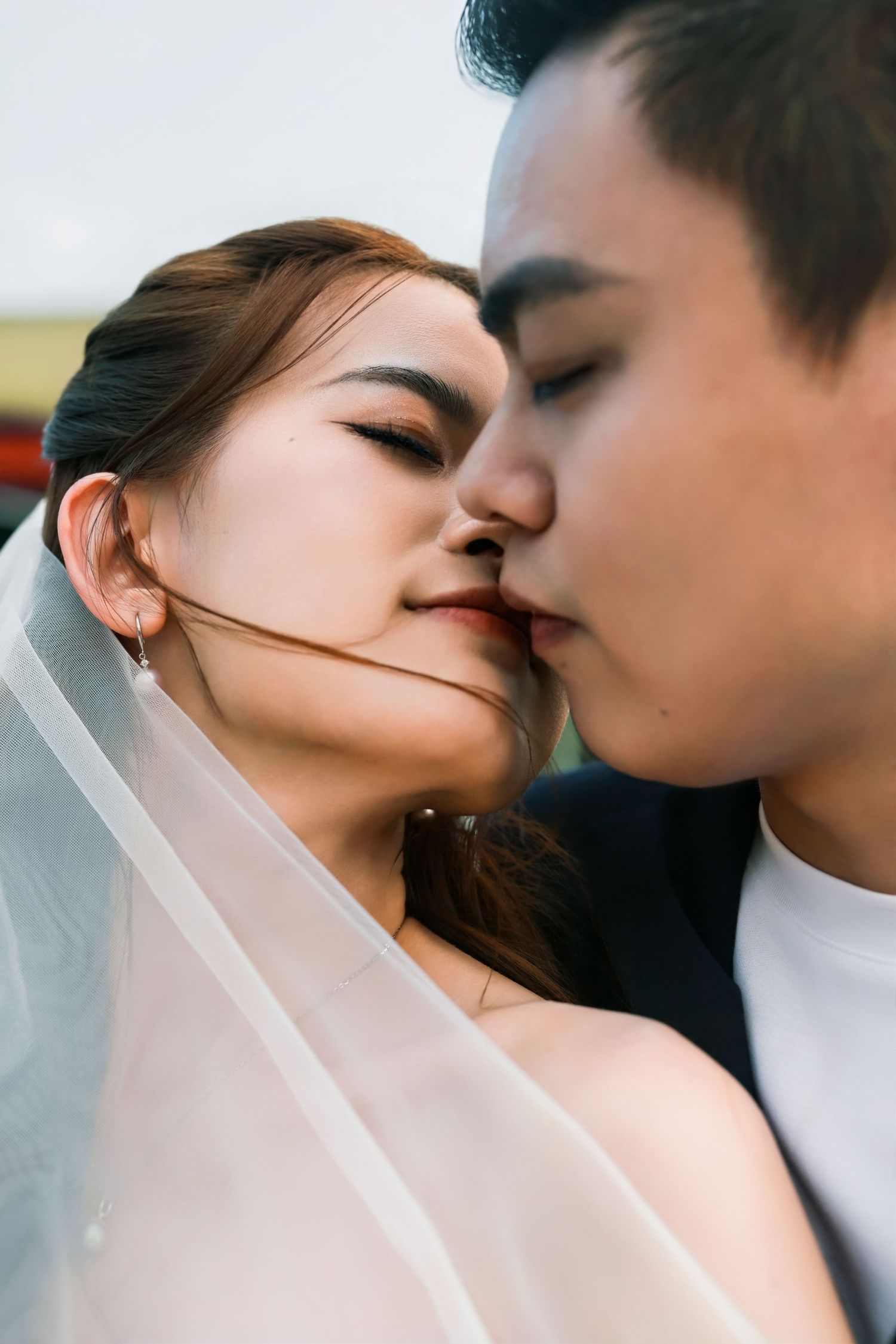 Asian couple sharing a romantic kiss during wedding, bride wearing veil and earrings in close-up portrait.