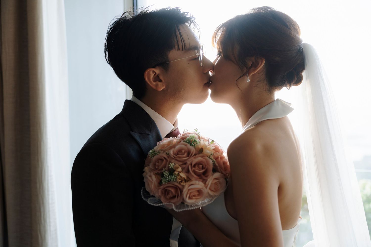 A bride and groom share a romantic kiss near a window, with the bride holding a bouquet of pink roses on their wedding day.