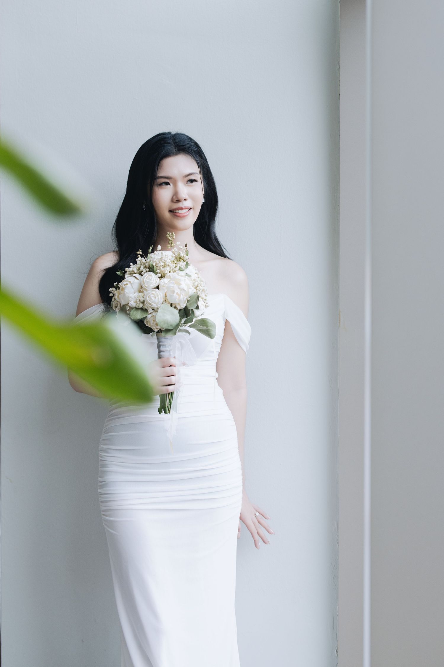 Elegant bride in white off-shoulder gown holding a bouquet of white roses and flowers, smiling softly indoors.