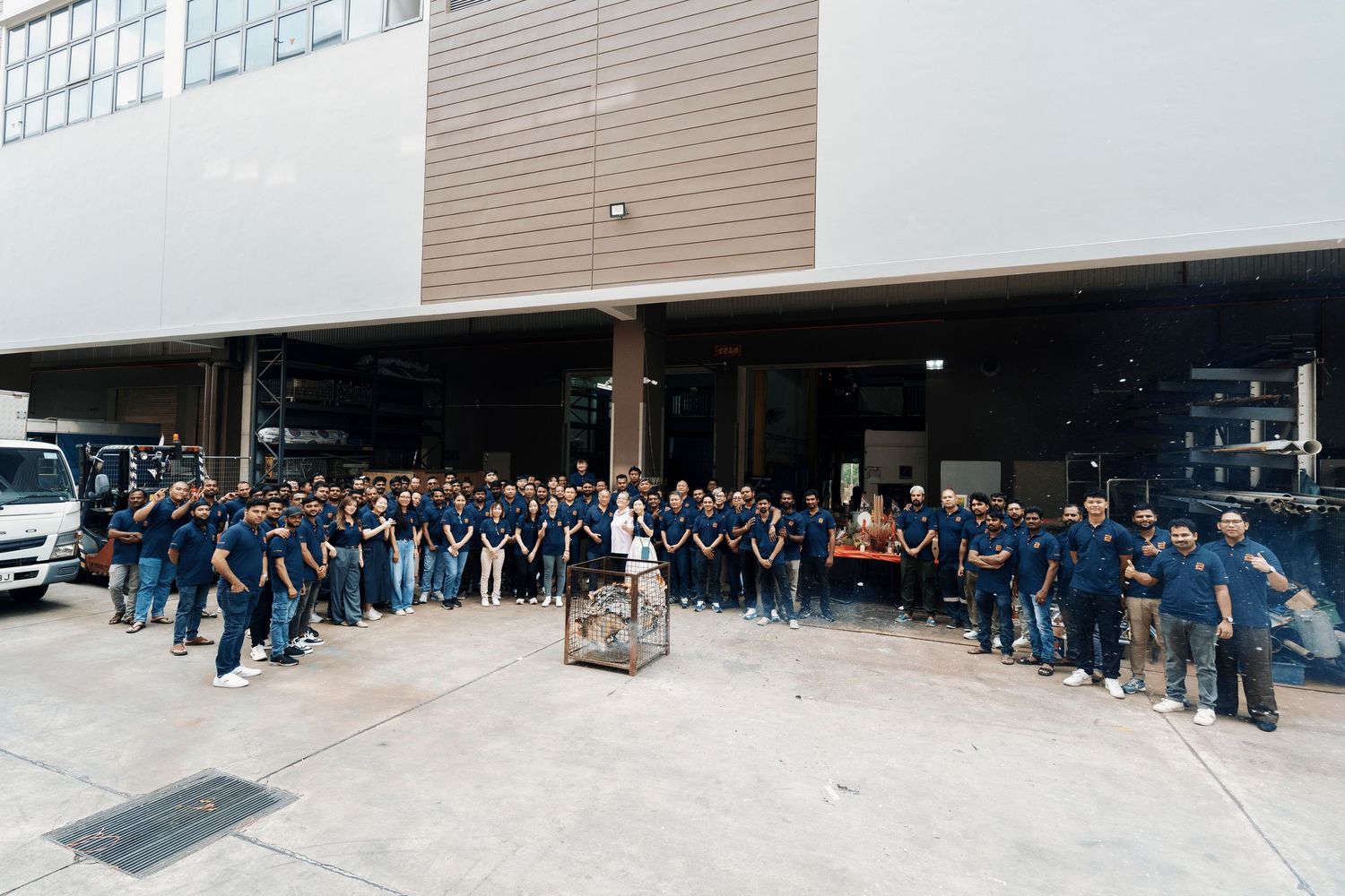Large group of factory workers in blue uniforms posing together outside an industrial warehouse facility.