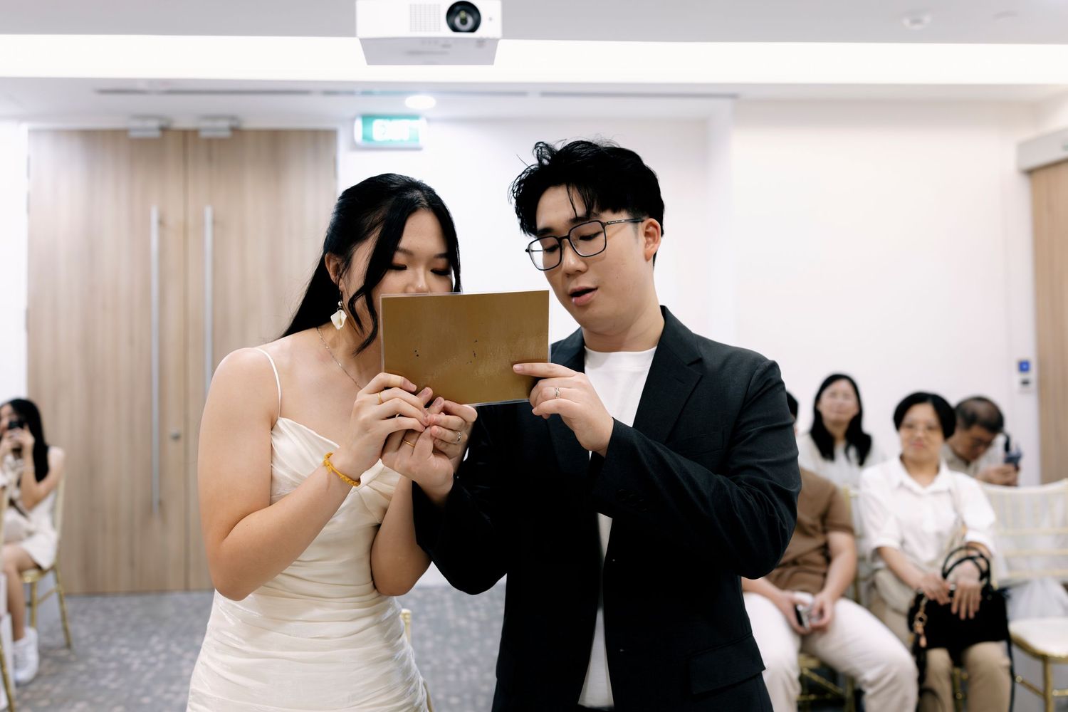 Couple reading vows from a golden card during an intimate wedding ceremony with guests seated in background.