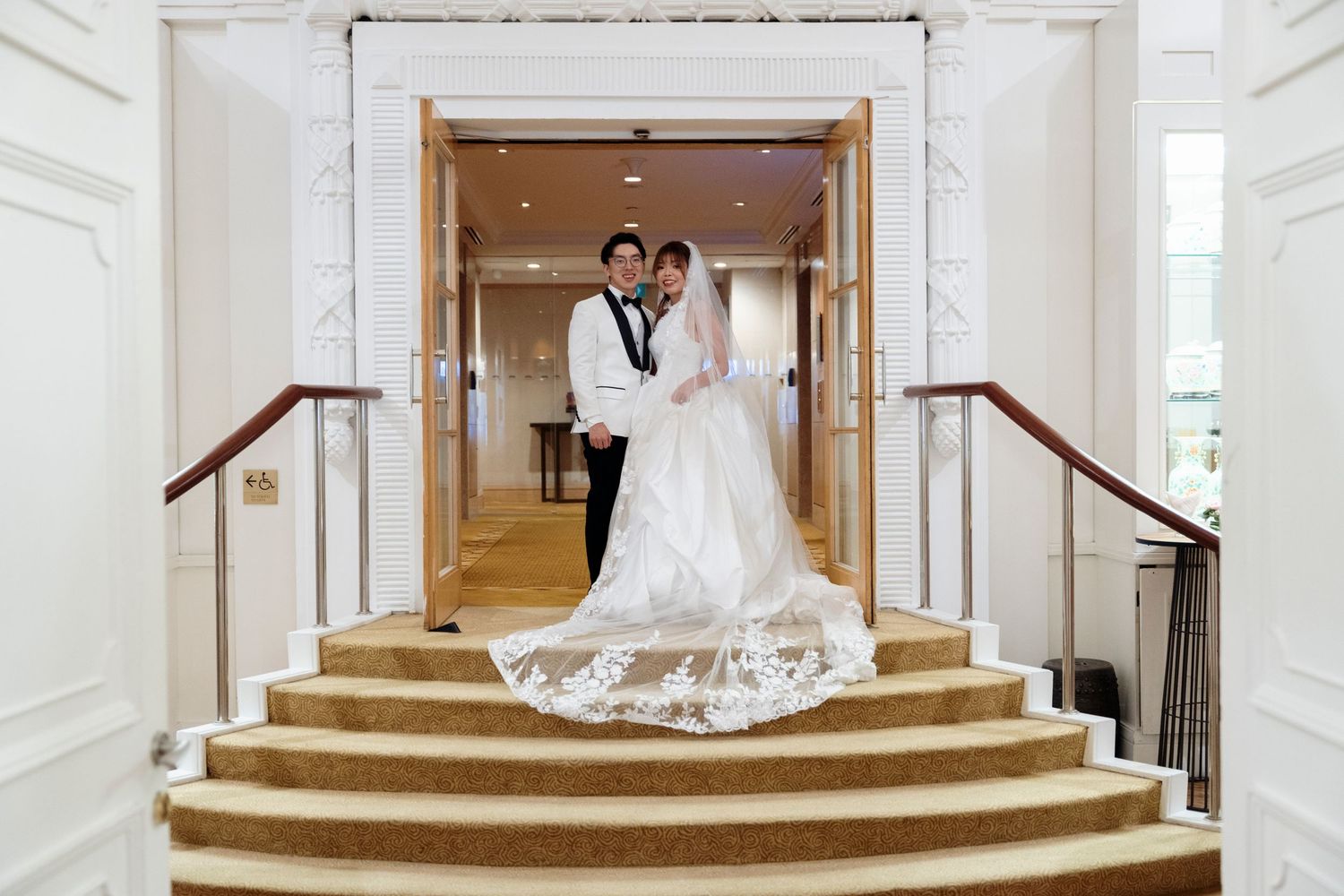 Bride in a white gown with a long lace train and groom in a tuxedo pose together at the top of a grand carpeted staircase.