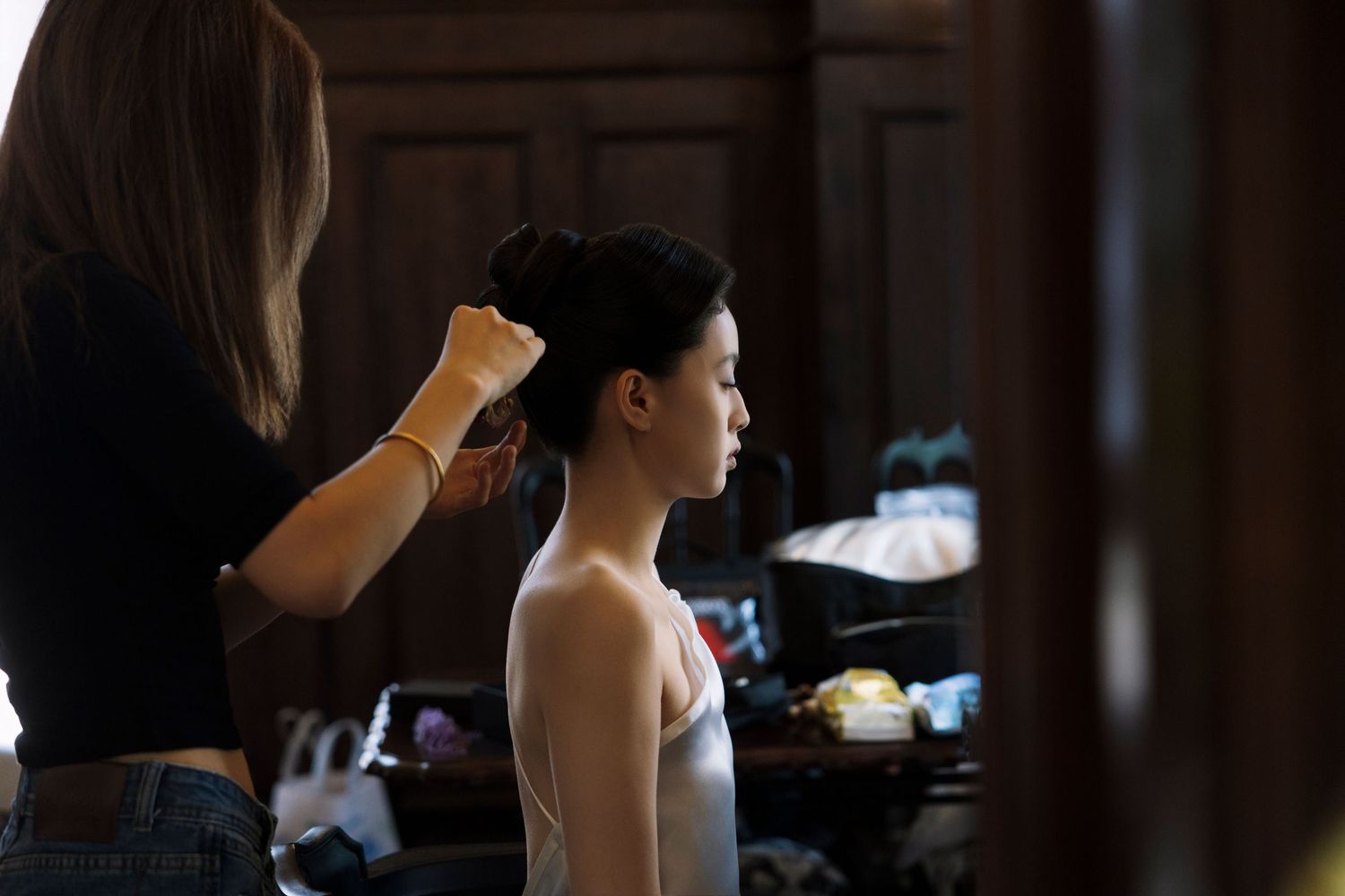 A hairstylist pins up a young woman's hair into a neat bun in a dimly lit backstage dressing room with wood paneling.