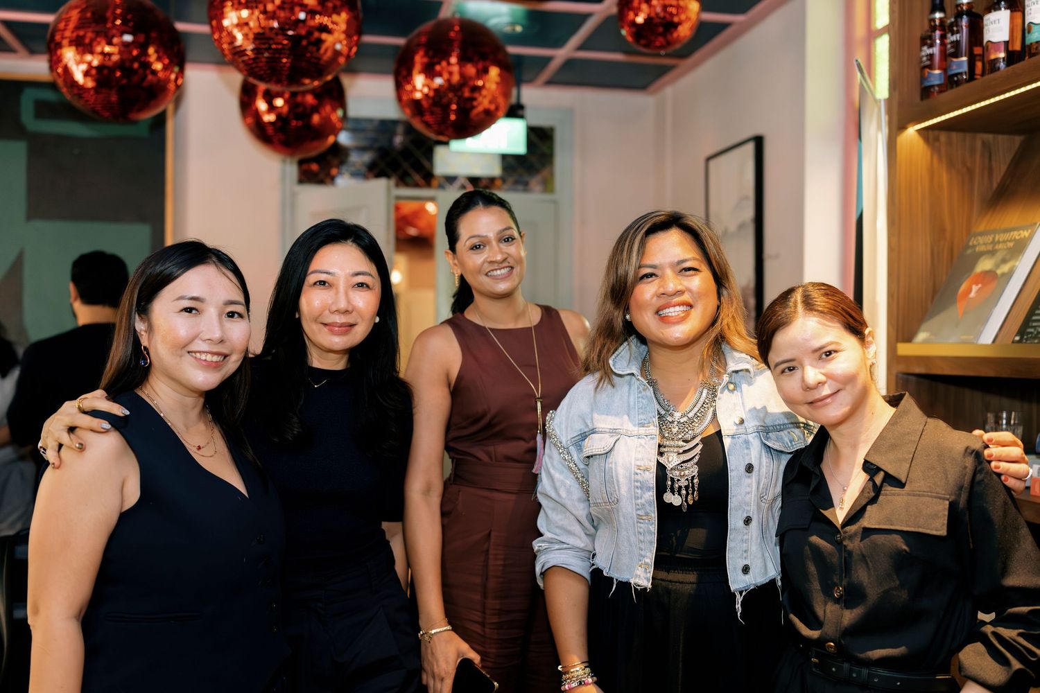 Five women smile together at an indoor social event decorated with red disco ball ornaments.