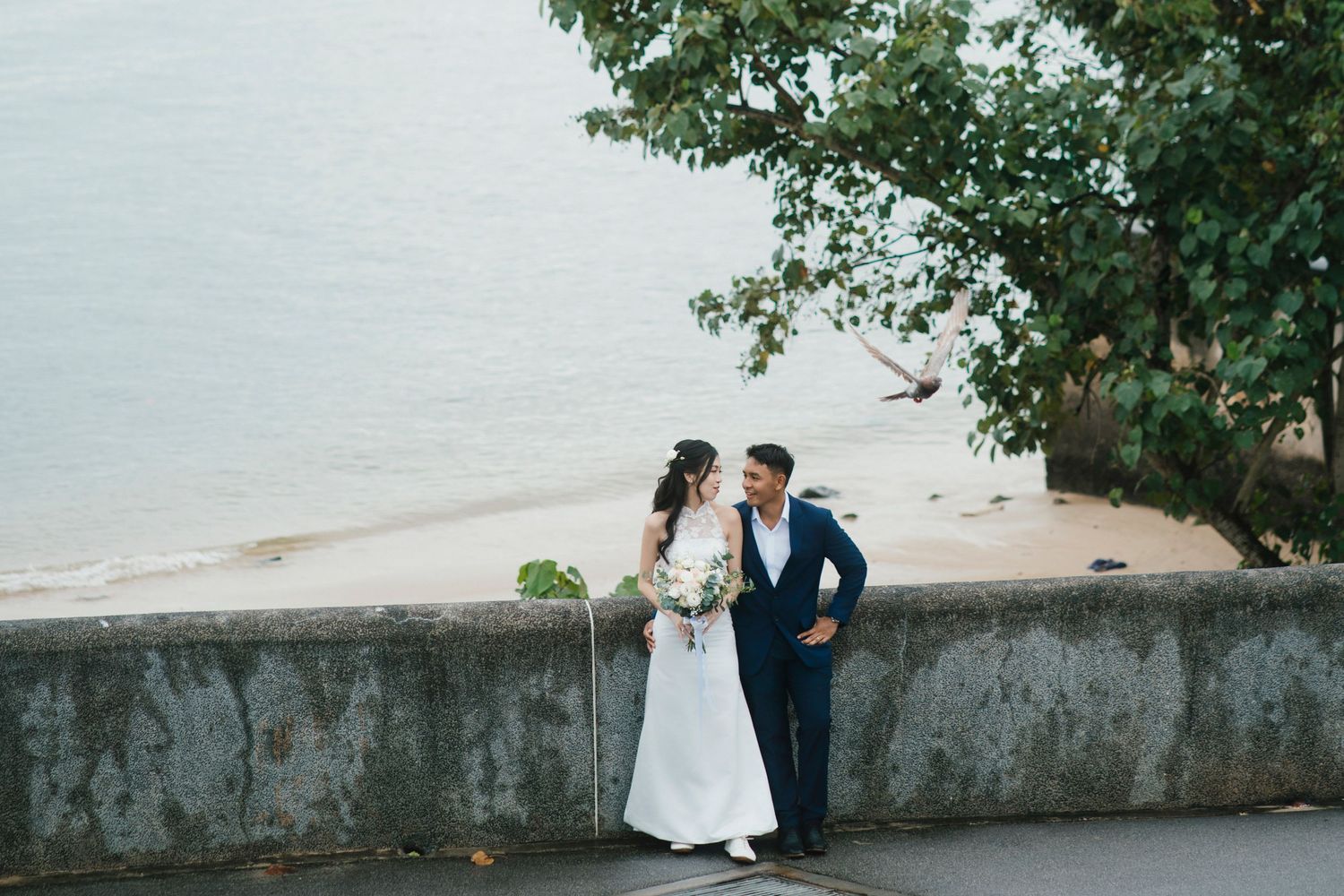 Bride and groom share a romantic moment by a seaside wall with a calm beach and tree in the background.
