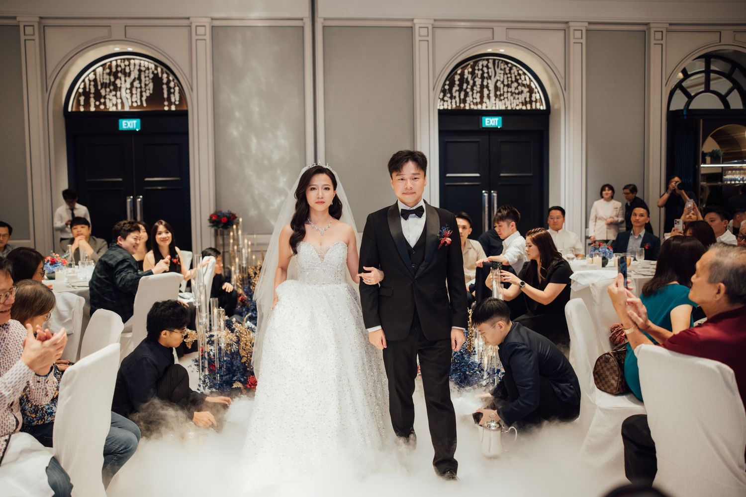 Bride in a white gown and groom in a black tuxedo walk arm in arm through a misty wedding reception hall as seated guests look on.