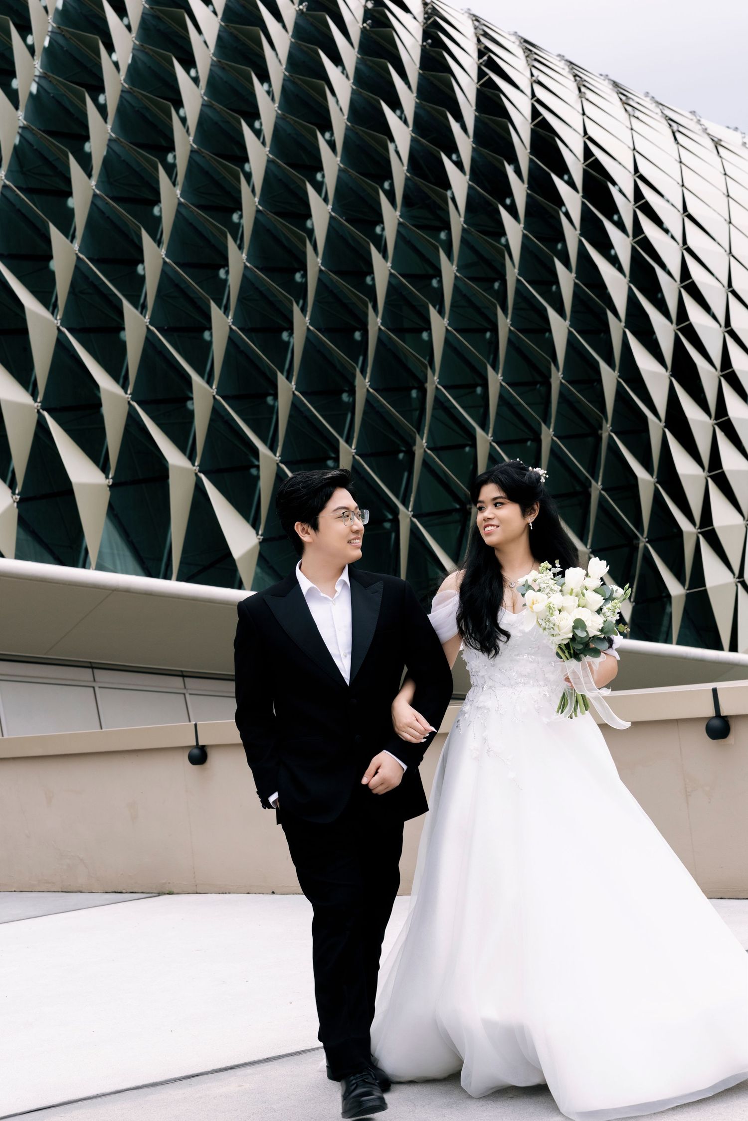 A couple in wedding attire walks arm in arm before a modern geometric building facade.