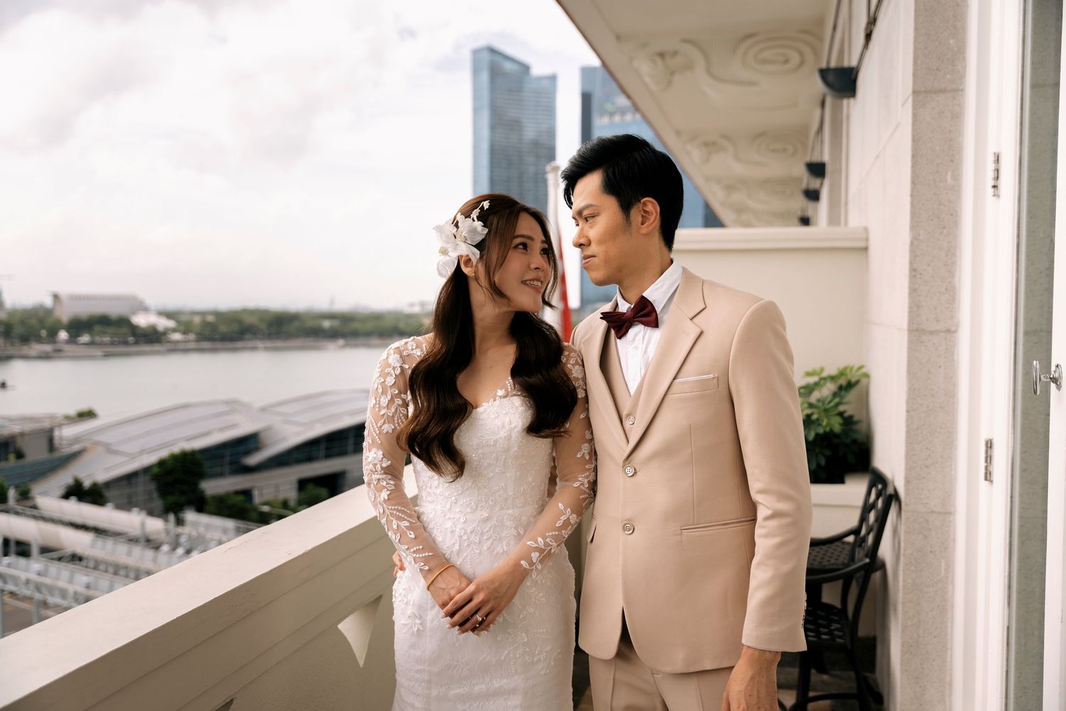 Couple in wedding attire on a balcony overlooking a waterfront cityscape, bride in lace gown and groom in beige suit.