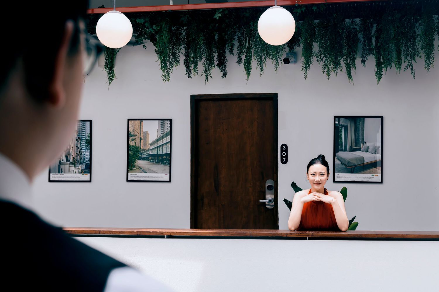 A smiling female hotel receptionist stands behind a wooden front desk in a modern, plant-adorned lobby.