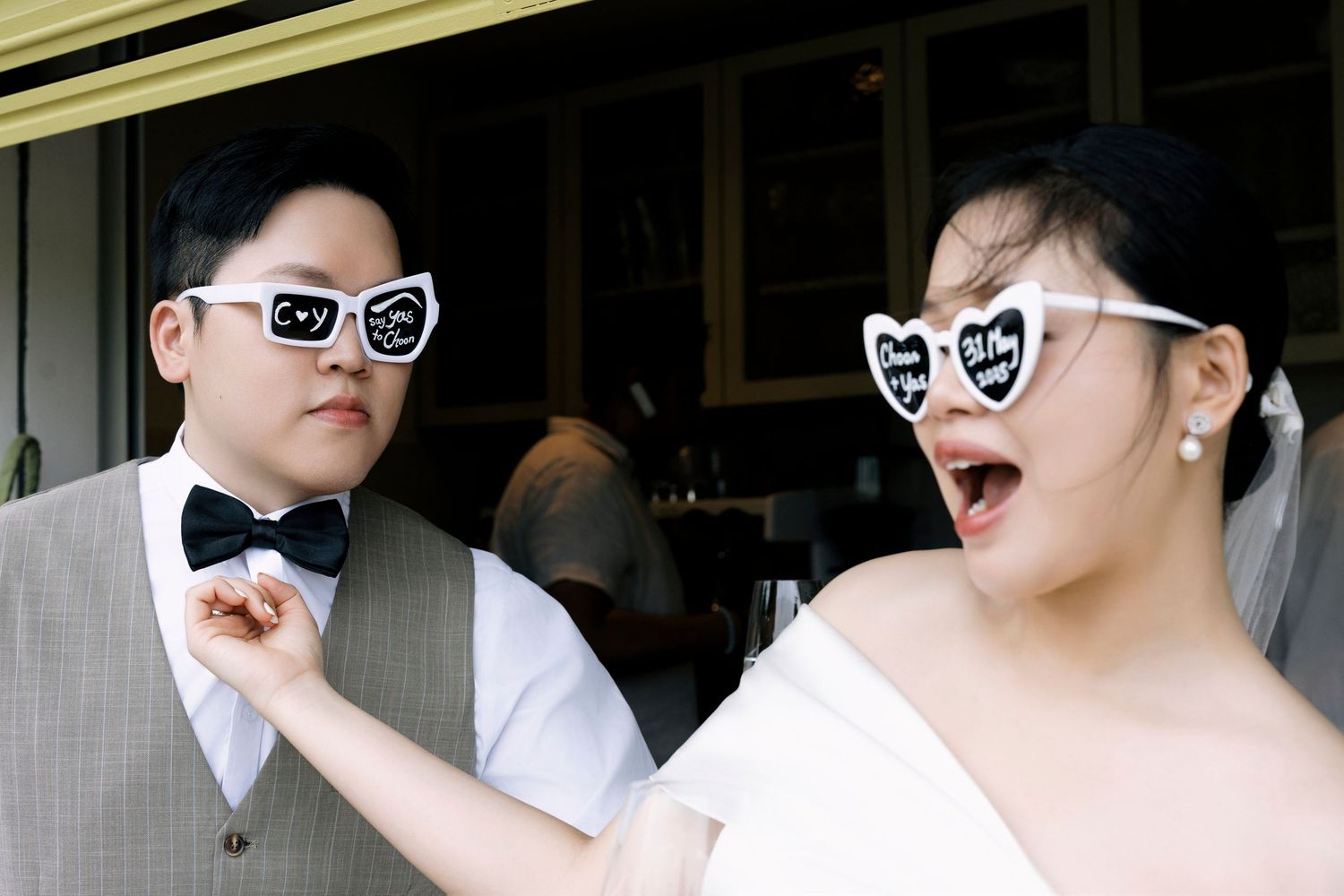 Playful Asian wedding couple wearing novelty heart-shaped sunglasses and laughing together outdoors.