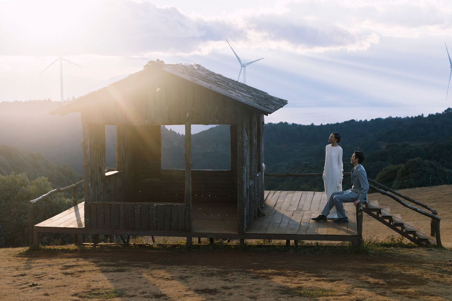 Couple in wedding attire standing on a rustic wooden deck with a wind turbine and golden sunset in the background.
