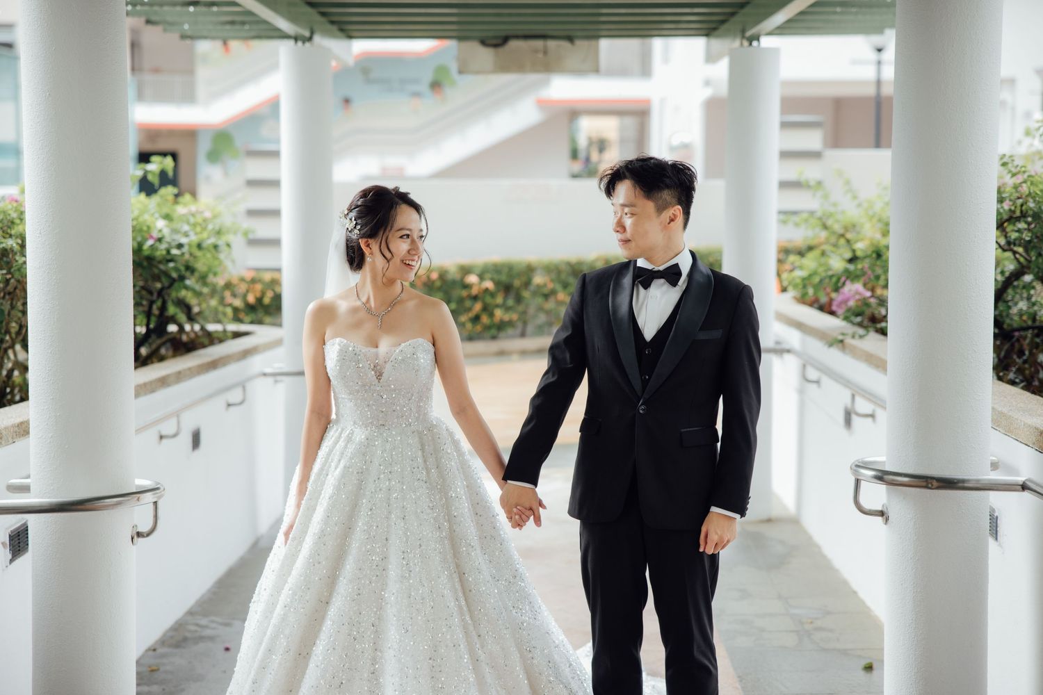 Bride in a white lace gown and groom in a black tuxedo with bow tie hold hands while walking between white columns on their wedding day.