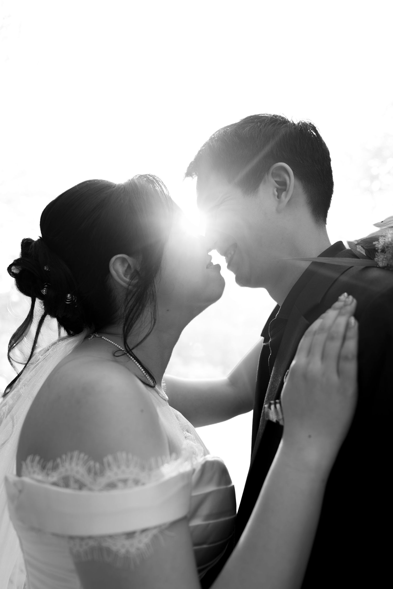 Black and white photo of a bride and groom sharing a romantic moment, backlit by sunlight, in wedding attire.