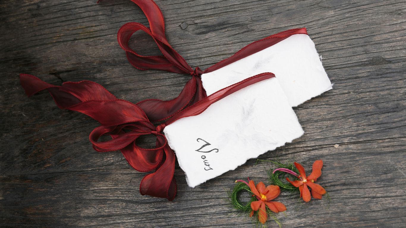A handwritten note card tied with dark red ribbon beside orange tropical flowers on rustic wooden surface.