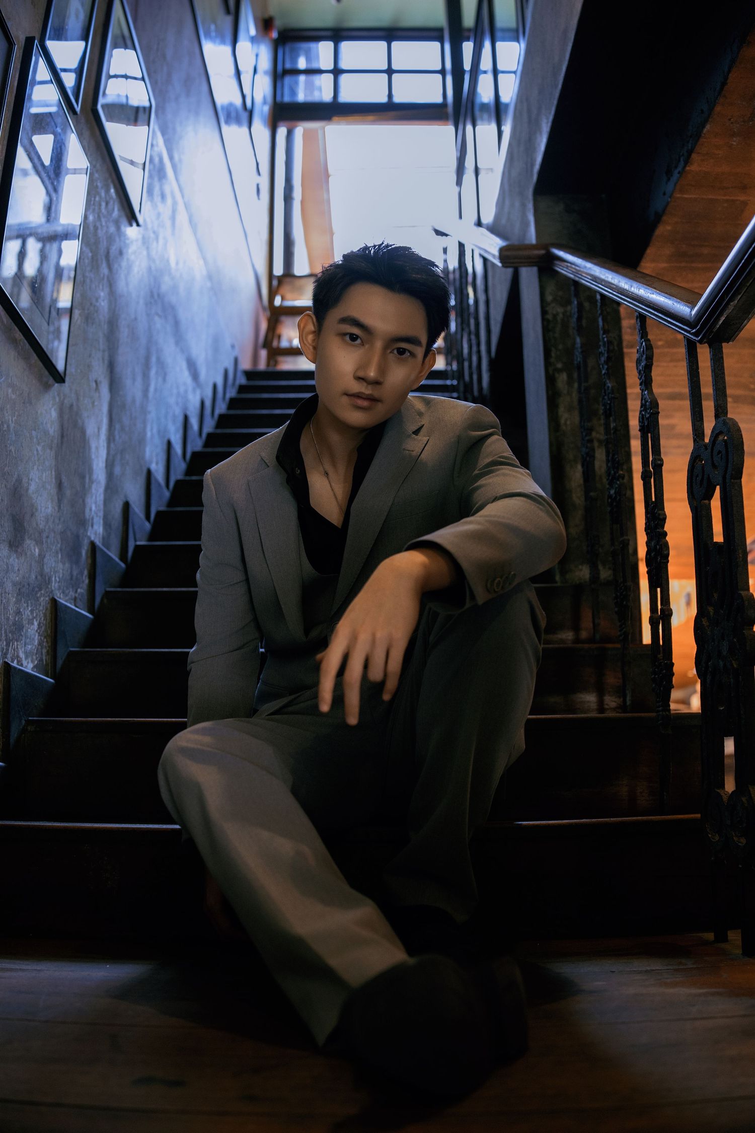 Stylish young Asian man in a gray suit poses confidently on dark interior staircase in dramatic lighting.