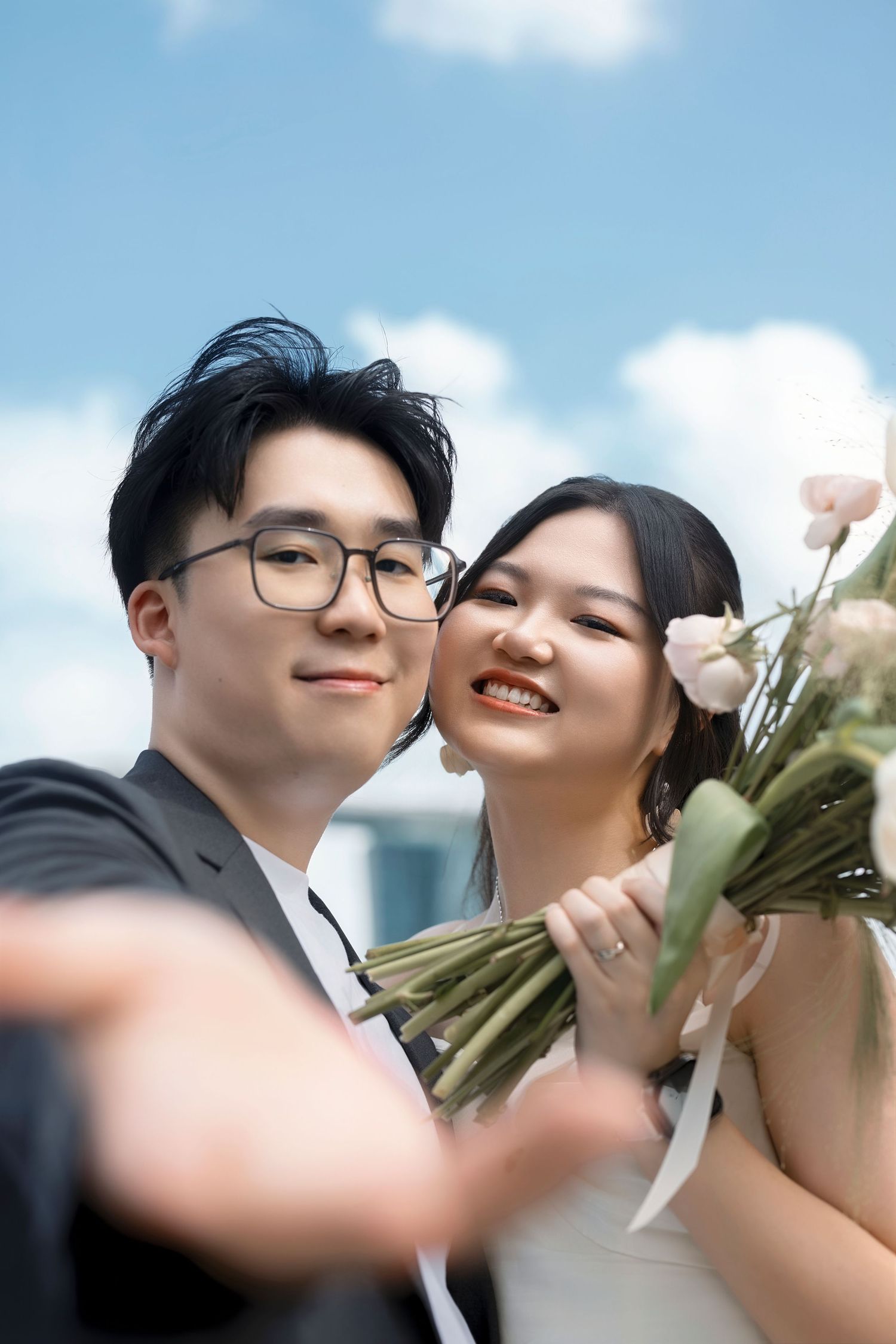 Smiling Asian couple in wedding attire pose for a selfie outdoors under a bright blue sky with clouds.