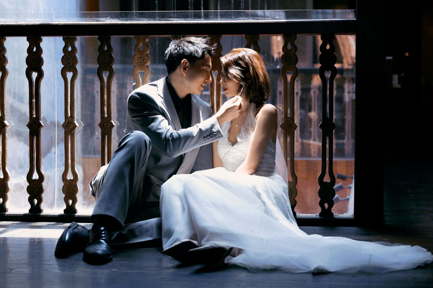 Romantic wedding couple sitting on balcony floor, groom in gray suit tenderly touching bride's face.