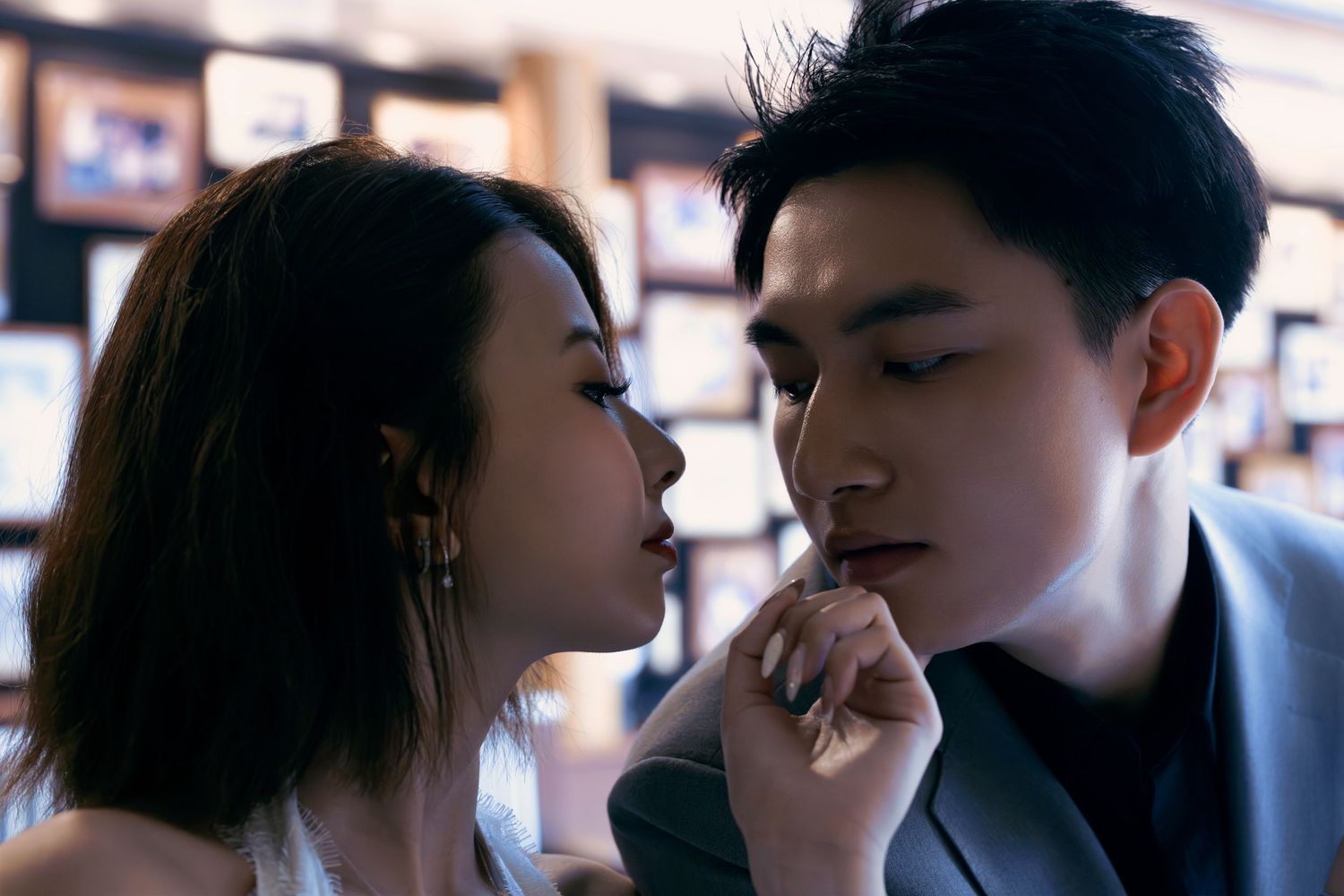 A couple shares a flirtatious close moment, the woman touching the mans chin in a warmly lit interior.