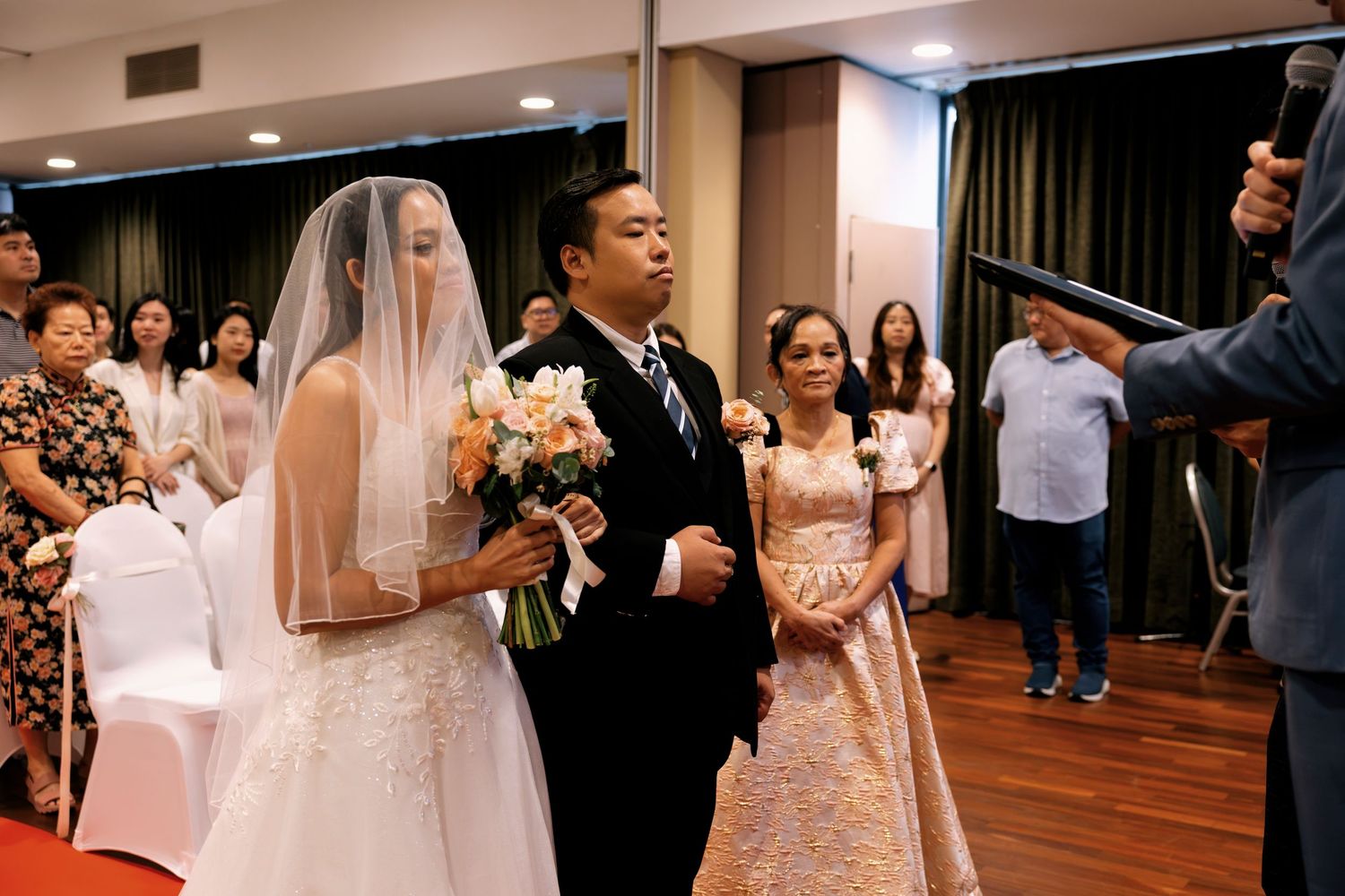 Bride in white gown and veil holding bouquet walks with groom during indoor wedding ceremony.
